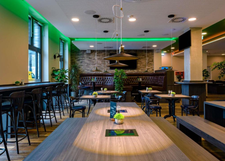 Moderne Hotelbar mit stilvoller Holzbestuhlung, grüner Akzentbeleuchtung und gemütlicher Lounge-Ecke, umgeben von Pflanzen, bietet einen einladenden Raum für entspannte Abende und geselliges Beisammensein.