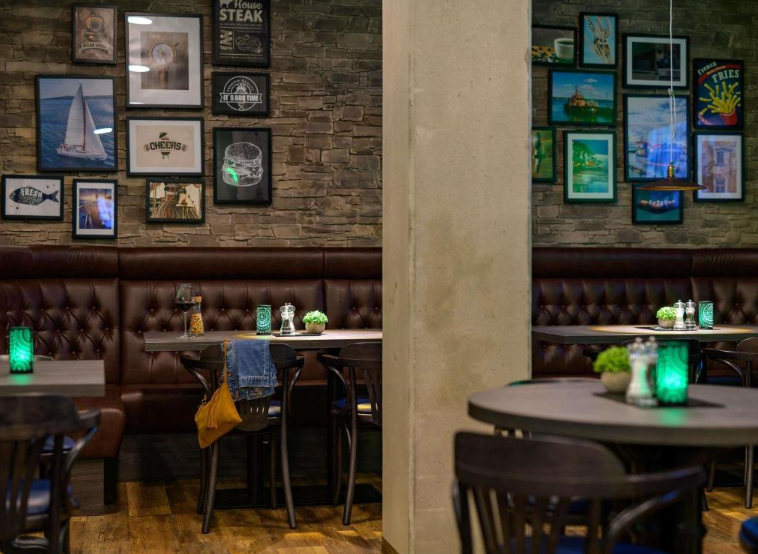 Gemütlicher Restaurantbereich mit braunen Ledersesseln, Holzstühlen und Tischen, dekoriert mit grünen Teelichtern. An den Wänden hängen zahlreiche bunte Bilder und Schwarz-Weiß-Fotografien, die eine einladende und entspannte Atmosphäre schaffen.