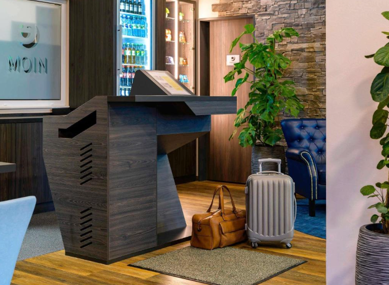 Hotelrezeption mit modernem Empfangstresen, Sitzbereich mit Sessel, Gepäckstücke am Boden und einem beleuchteten Getränke- und Snackangebot im Hintergrund, gemütliche Atmosphäre mit Pflanzen und Holzboden.
