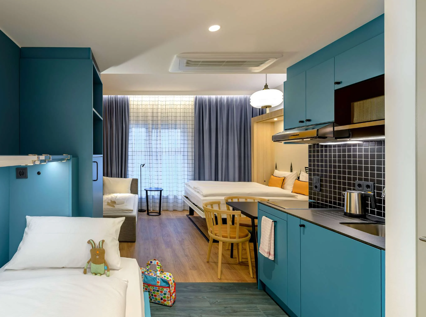 Moderner Hotelzimmerbereich mit integrierter Küchenzeile, Essbereich und Schlafgelegenheiten. Helle, freundliche Atmosphäre mit Holz- und Blautönen, großen Fenstern und Vorhängen für viel Tageslicht und Gemütlichkeit. Ideal für längere Aufenthalte und Familien oder Geschäftsreisende.