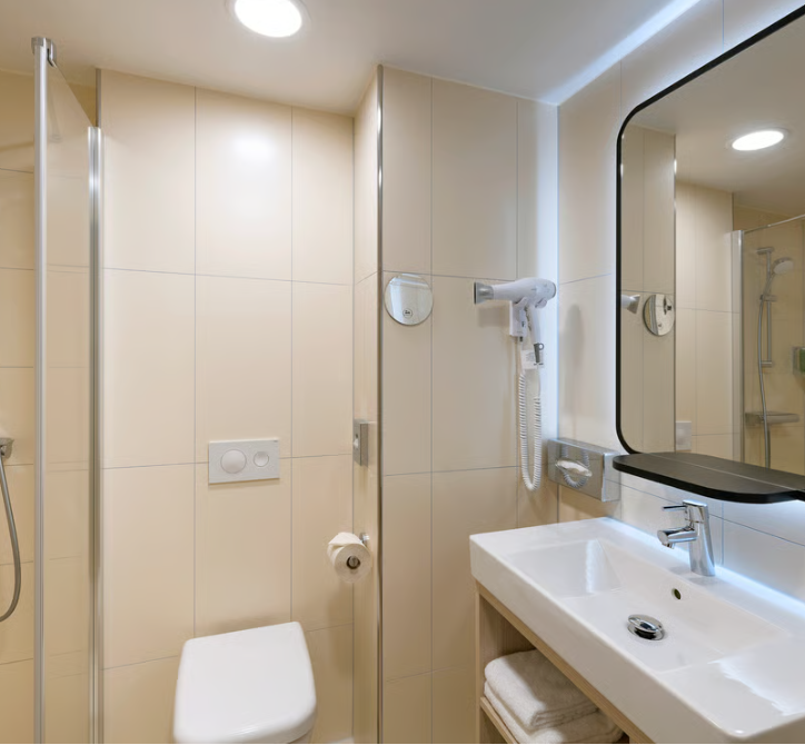 Kompaktes, modernes Badezimmer mit weißer Toilette, großem Spiegel, integriertem Waschbecken, Haartrockner an der Wand und offener Glasdusche. Helle, beige Fliesen schaffen eine freundliche Atmosphäre.