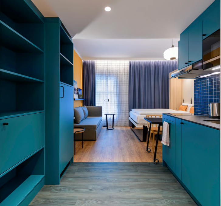 Modern eingerichtetes Hotelzimmer mit Kochbereich, Sitzecke, Bett und großem Fenster mit Vorhängen, das eine gemütliche und funktionale Atmosphäre schafft.