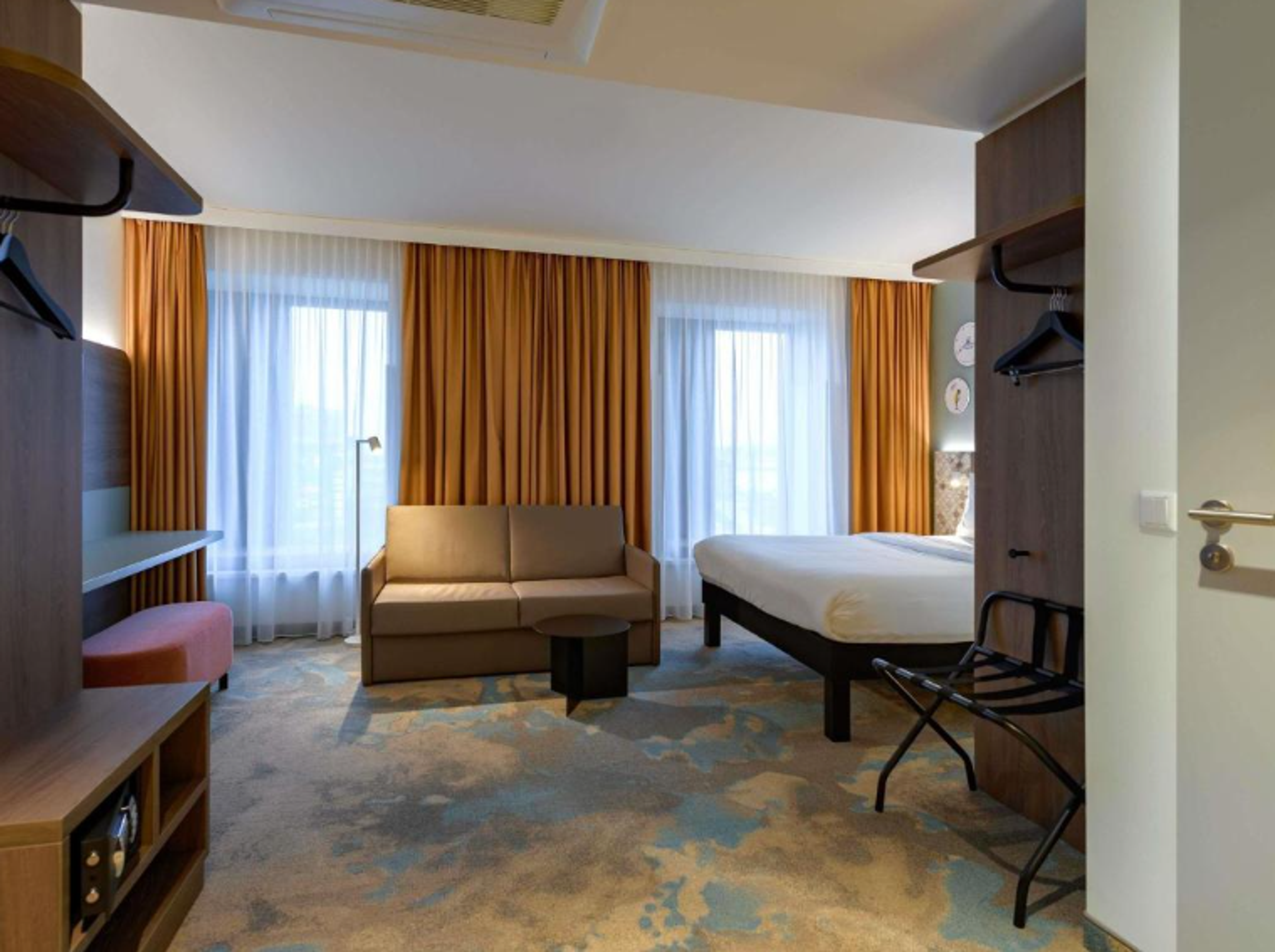Gemütliches Hotelzimmer mit großem Bett, Sofa, modernen Möbeln und warmen Vorhängen. Helle Raumgestaltung mit Tageslicht durch zwei Fenster, einladende Atmosphäre für Erholung und Komfort. Gemütliches Hotelzimmer mit großem Bett, Sofa, modernen Möbeln und warmen Vorhängen. Helle Raumgestaltung mit Tageslicht durch zwei Fenster, einladende Atmosphäre für Erholung und Komfort.