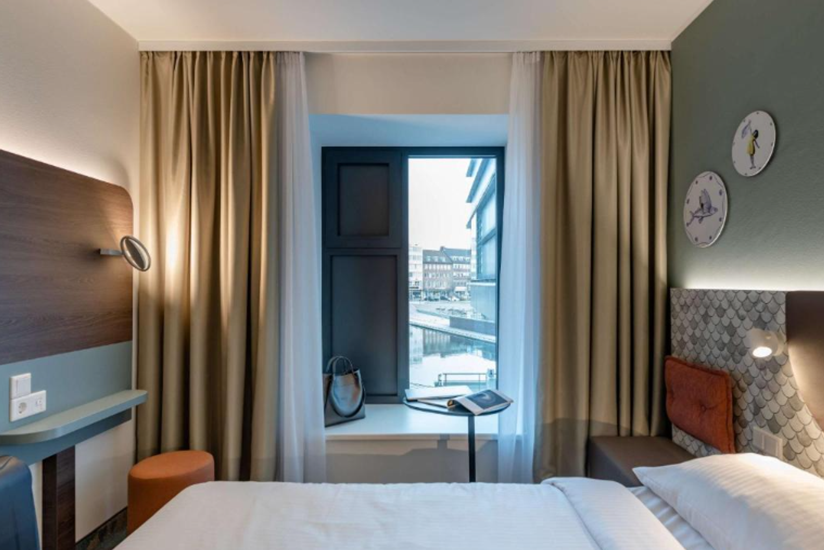 Modernes Hotelzimmer mit gemütlichem Doppelbett, Hell-Dunkel-Kontrast, Fenster mit Stadtblick und praktischem Arbeitsplatz am Fensterbank. Schlichte, elegante Einrichtung in warmen Farbtönen.