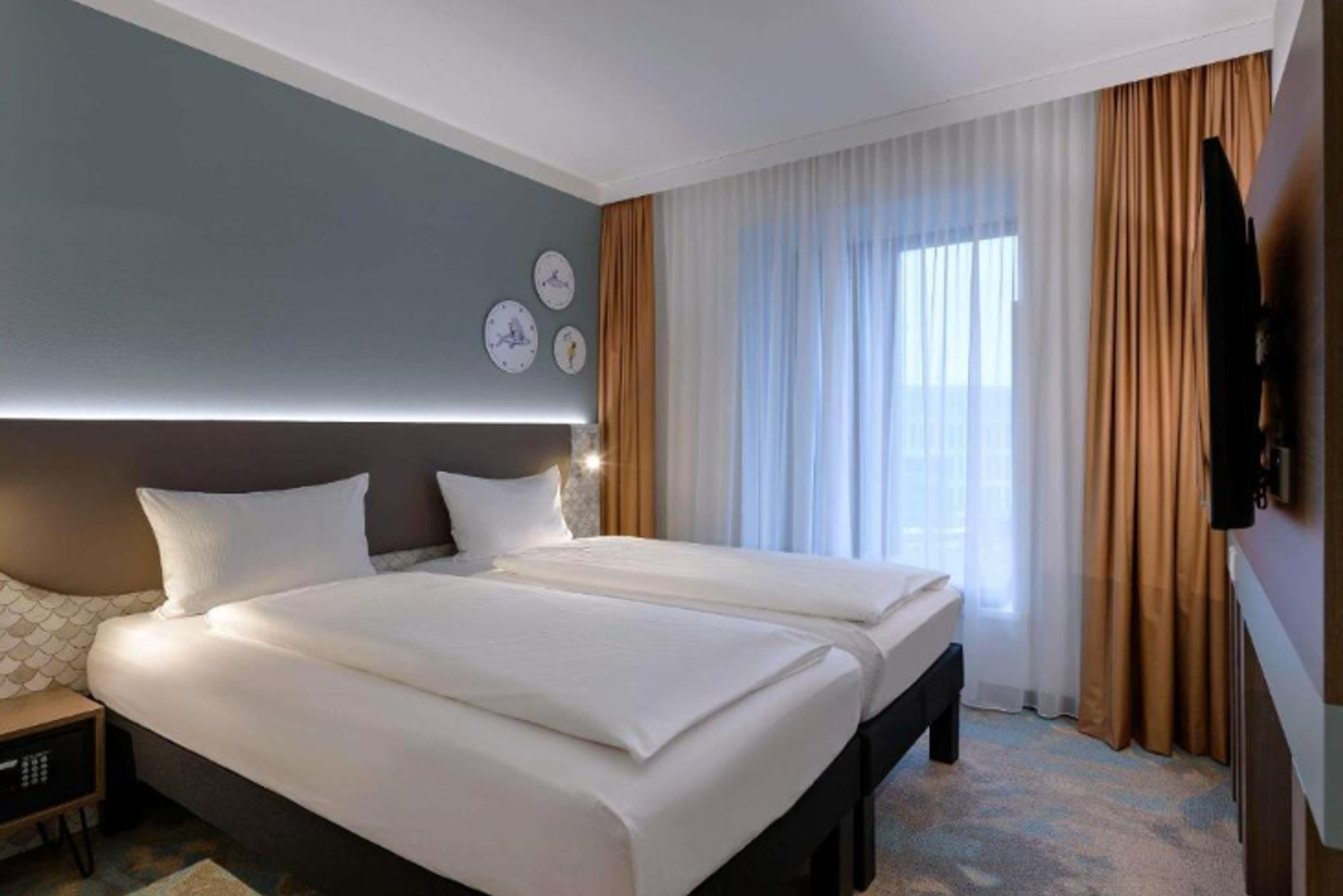 Modern eingerichtetes Doppelzimmer mit zwei Einzelbetten, großer Fensterfront mit Vorhängen, beruhigenden Farben und stilvollen Wanduhren für eine entspannte Atmosphäre.