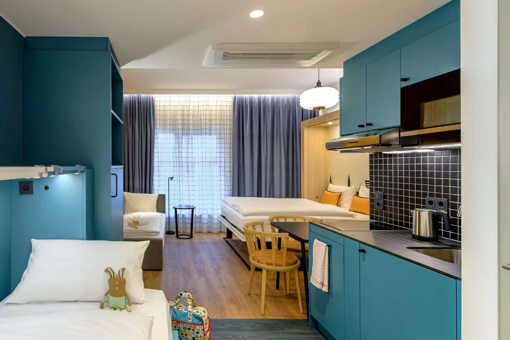 Modern eingerichtetes Zimmer mit blauen Möbeln, separatem Schlaf- und Wohnzimmerbereich, einer kleinen Küche und großem Fenster mit Tageslicht. Gemütliche Atmosphäre für komfortablen Aufenthalt.