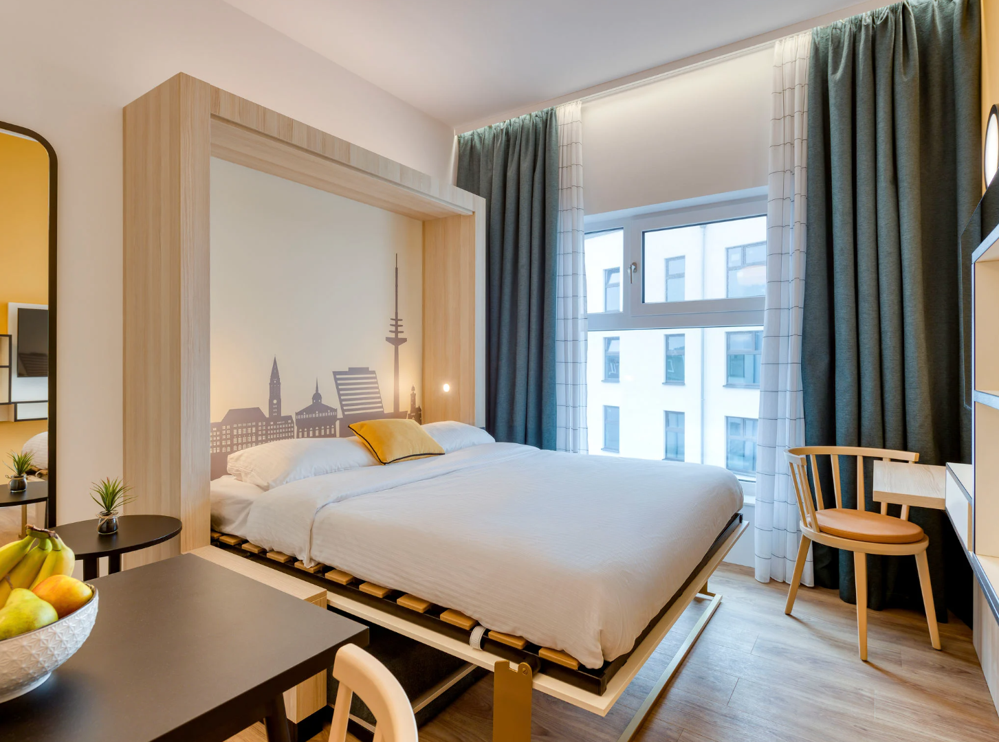 Helles, modernes Zimmer mit großem Bett, beruhigender Stadt-Silhouette an der Wand, großen Fenstern mit Tageslicht, gemütlichen Vorhängen und schlichter Holzmöblierung für komfortablen Aufenthalt.