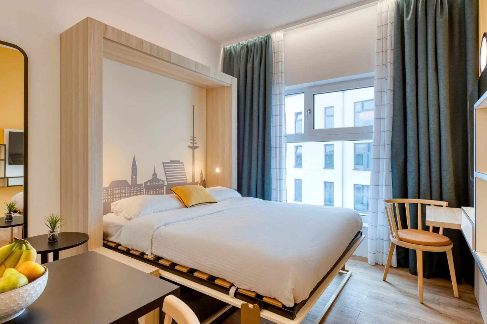 Helles, modernes Zimmer mit großem Bett, beruhigender Stadt-Silhouette an der Wand, großen Fenstern mit Tageslicht, gemütlichen Vorhängen und schlichter Holzmöblierung für komfortablen Aufenthalt.