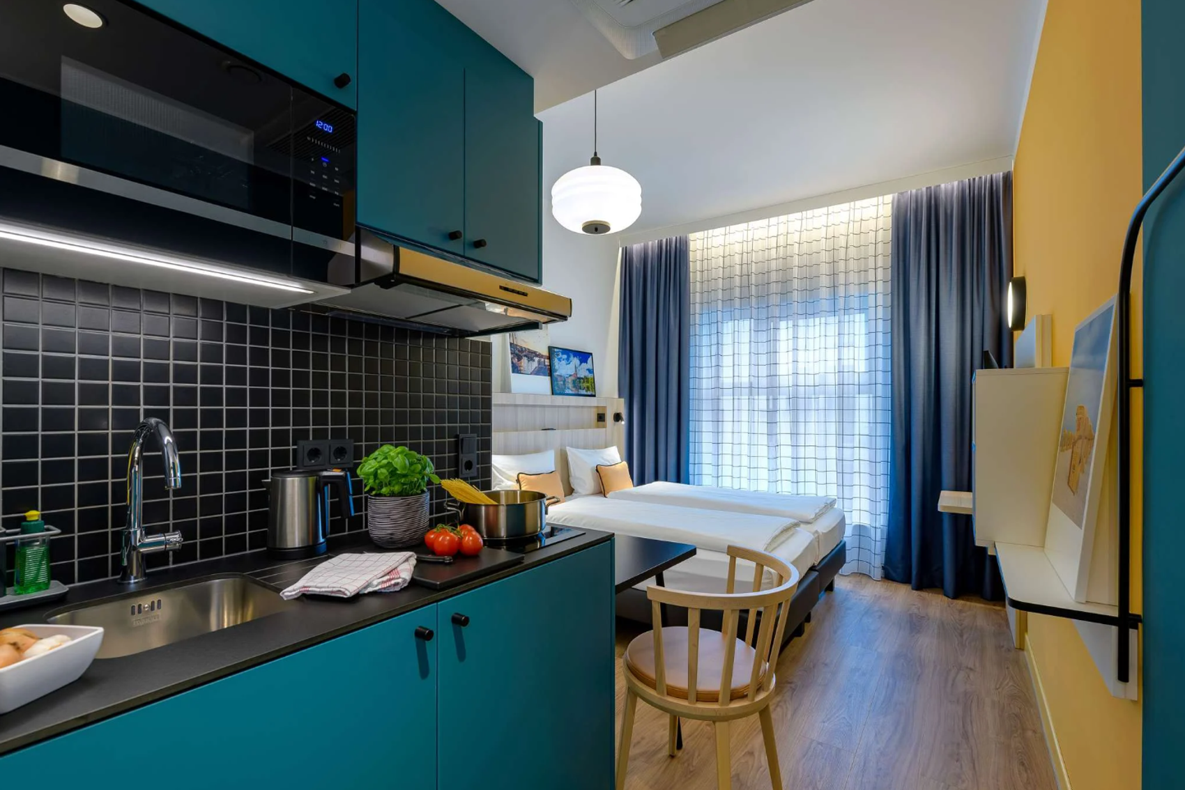 Modernes Hotelzimmer mit heller Atmosphäre, gemütlichem Doppelbett, kleiner Küchenzeile mit blauen Schränken und Küchenutensilien. Großes Fenster mit lichtdurchlässigen Vorhängen sorgt für freundlichen Raum. Holzfußboden und schlichte Möblierung verstärken das einladende Ambiente.