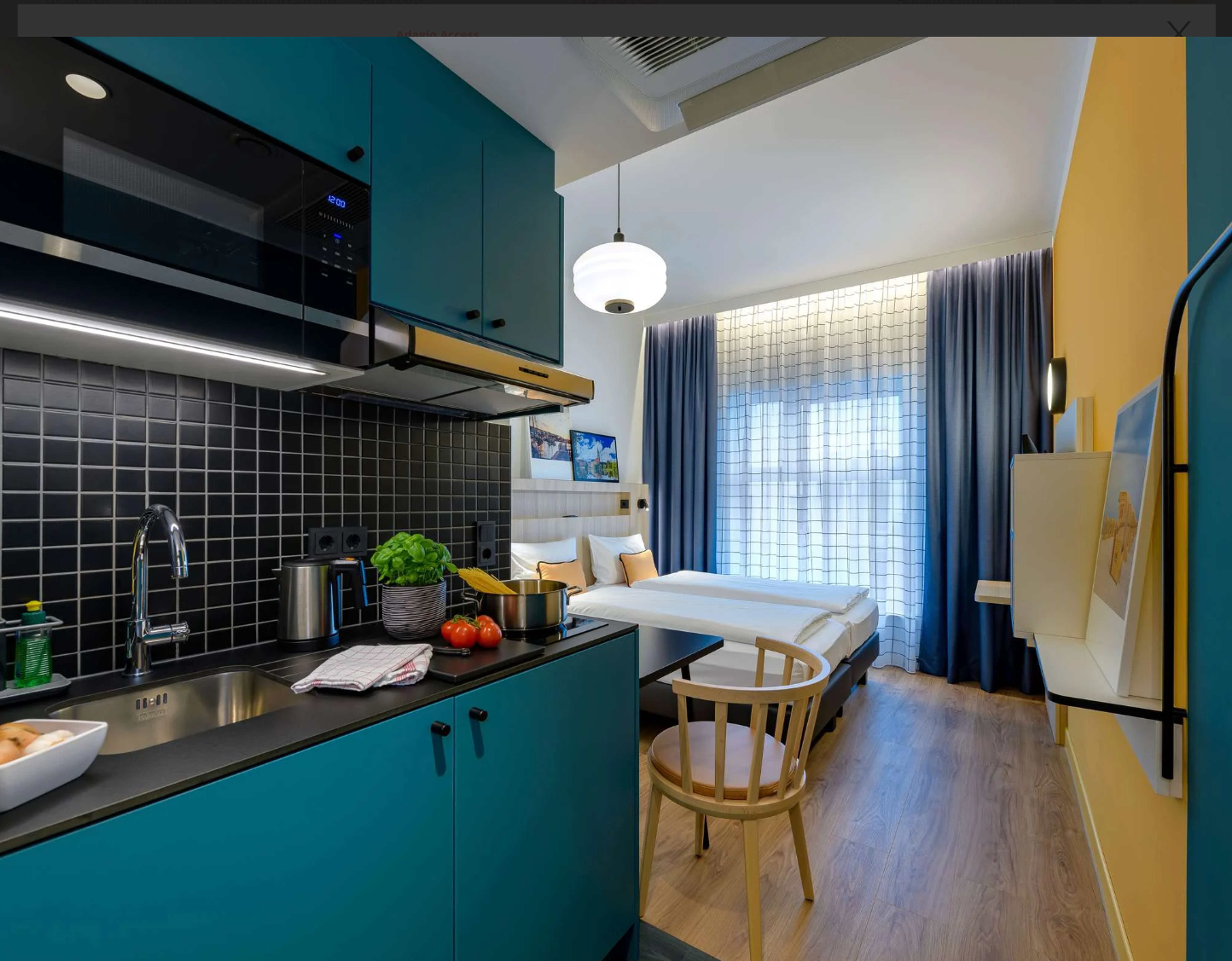 Modernes Hotelzimmer mit heller Atmosphäre, gemütlichem Doppelbett, kleiner Küchenzeile mit blauen Schränken und Küchenutensilien. Großes Fenster mit lichtdurchlässigen Vorhängen sorgt für freundlichen Raum. Holzfußboden und schlichte Möblierung verstärken das einladende Ambiente. Modernes Hotelzimmer mit heller Atmosphäre, gemütlichem Doppelbett, kleiner Küchenzeile mit blauen Schränken und Küchenutensilien. Großes Fenster mit lichtdurchlässigen Vorhängen sorgt für freundlichen Raum. Holzfußboden und schlichte Möblierung verstärken das einladende Ambiente.