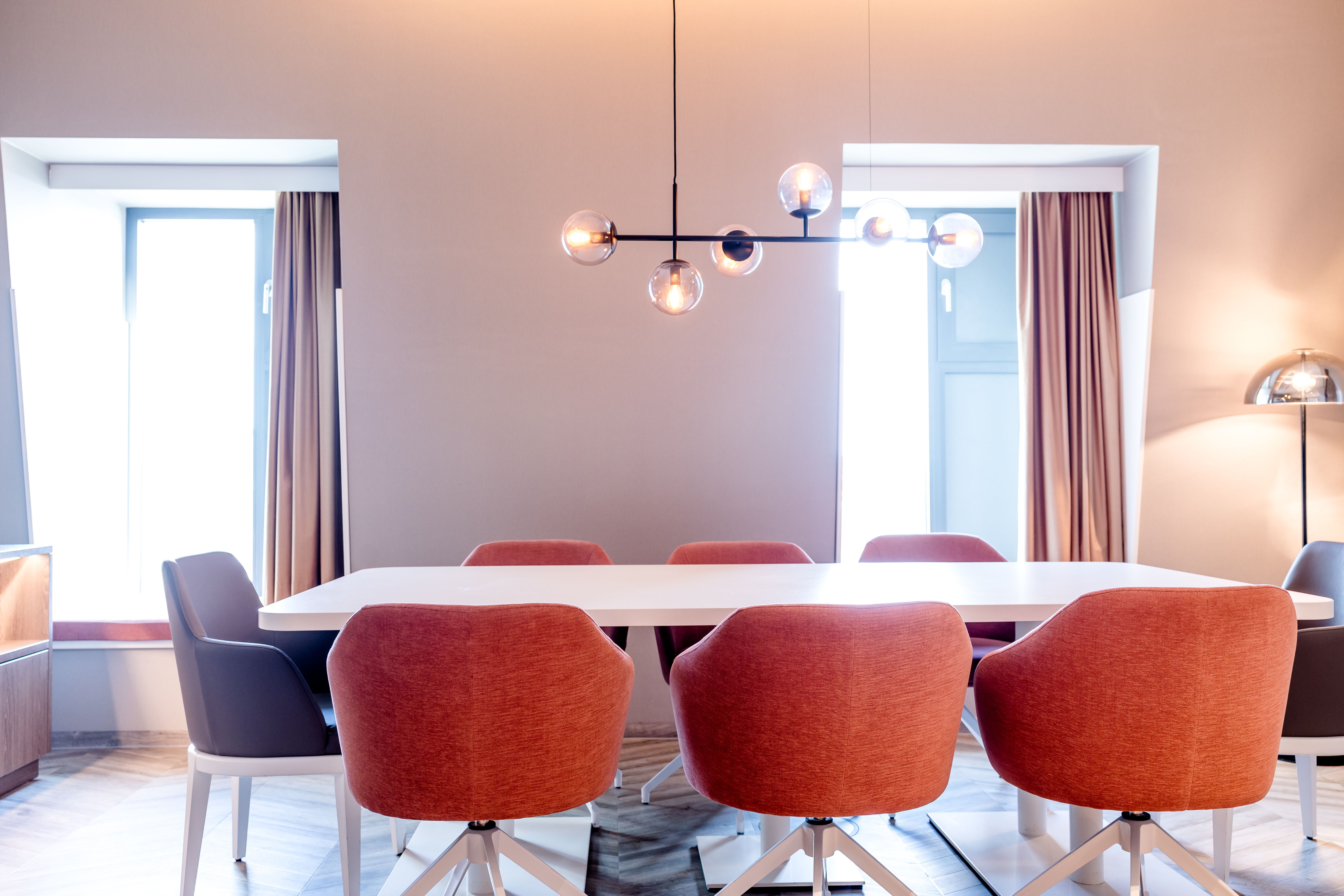 Moderner Konferenzraum mit großem weißem Tisch, acht bequemen, rot-orangefarbenen Stühlen und zwei Fenstern, die Tageslicht hereinlassen. Dezente Beleuchtung durch eine stilvolle Hängelampe und stehende Lampe schafft eine helle, einladende Atmosphäre.