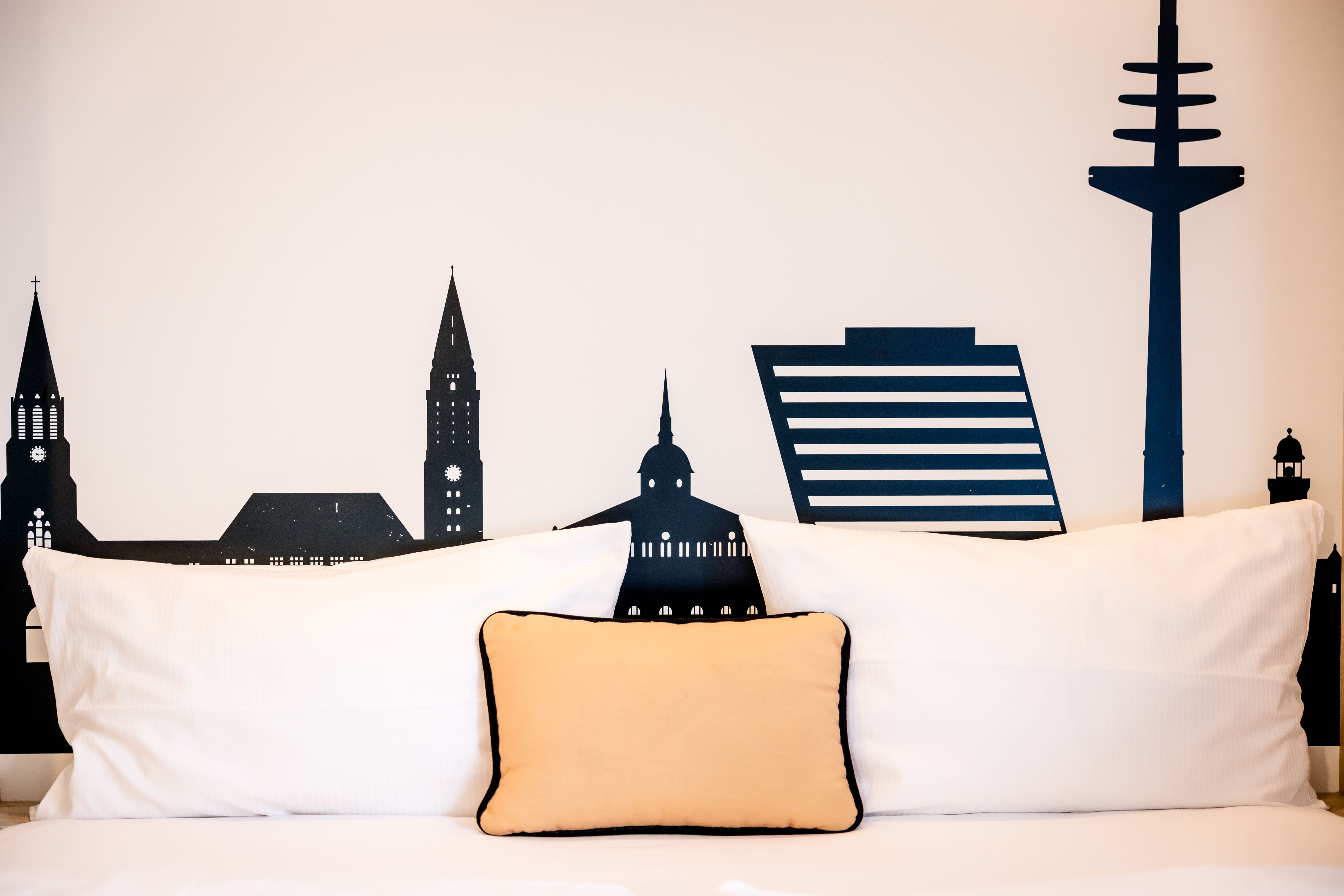 Kopfteil mit stilisierter Stadtsilhouette hinter einem großen Bett mit weißen Kissen und einem kleinen, beigen Kissen in der Mitte, vermittelt urbane Atmosphäre und modernes, gemütliches Ambiente.