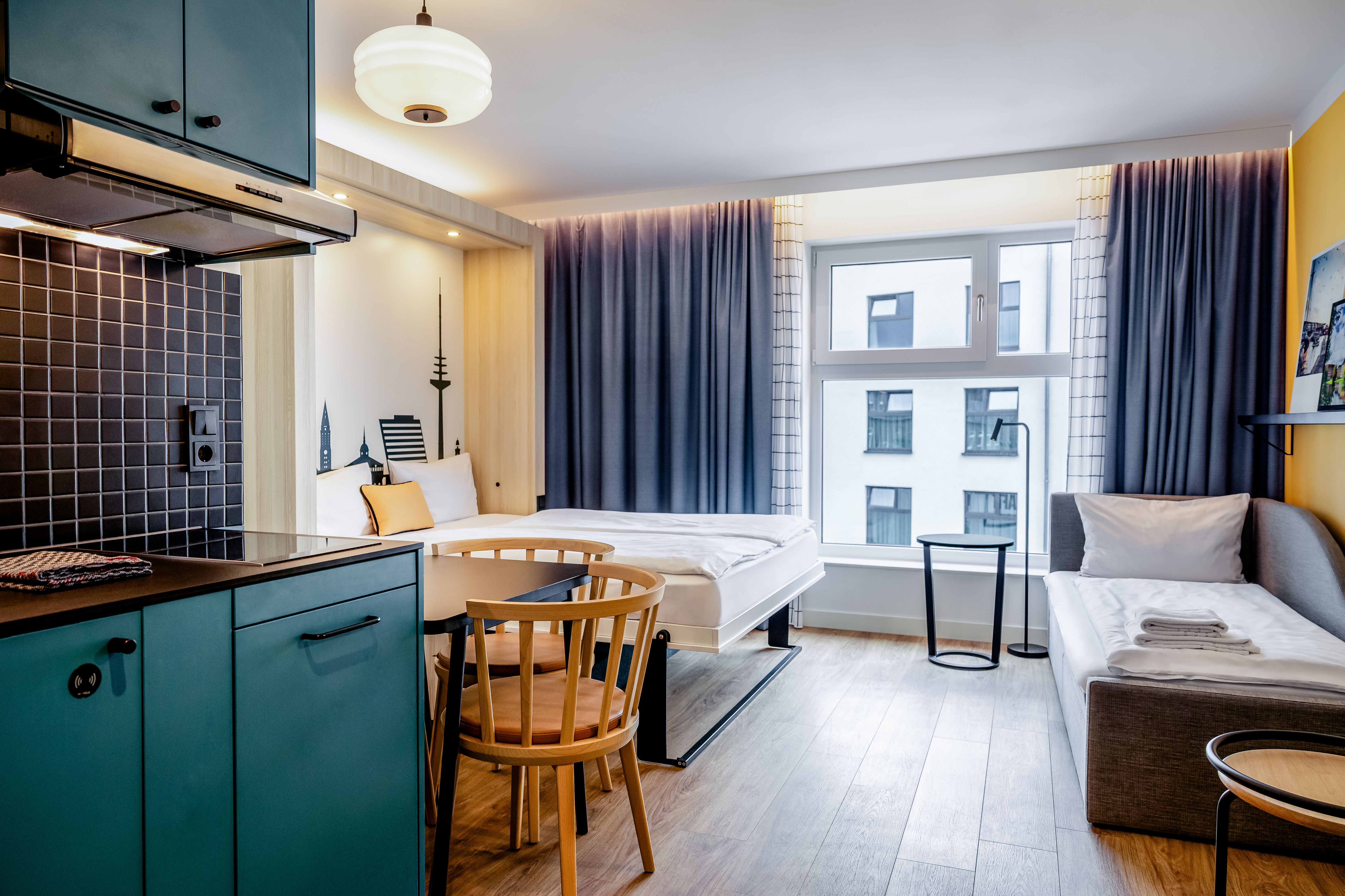Helles, modernes Studio-Apartment mit Kochzeile, Essbereich, zwei Einzelbetten, großem Fenster und Blick auf umliegende Gebäude. Warme Holz- und sanfte Blautöne schaffen eine einladende Atmosphäre.