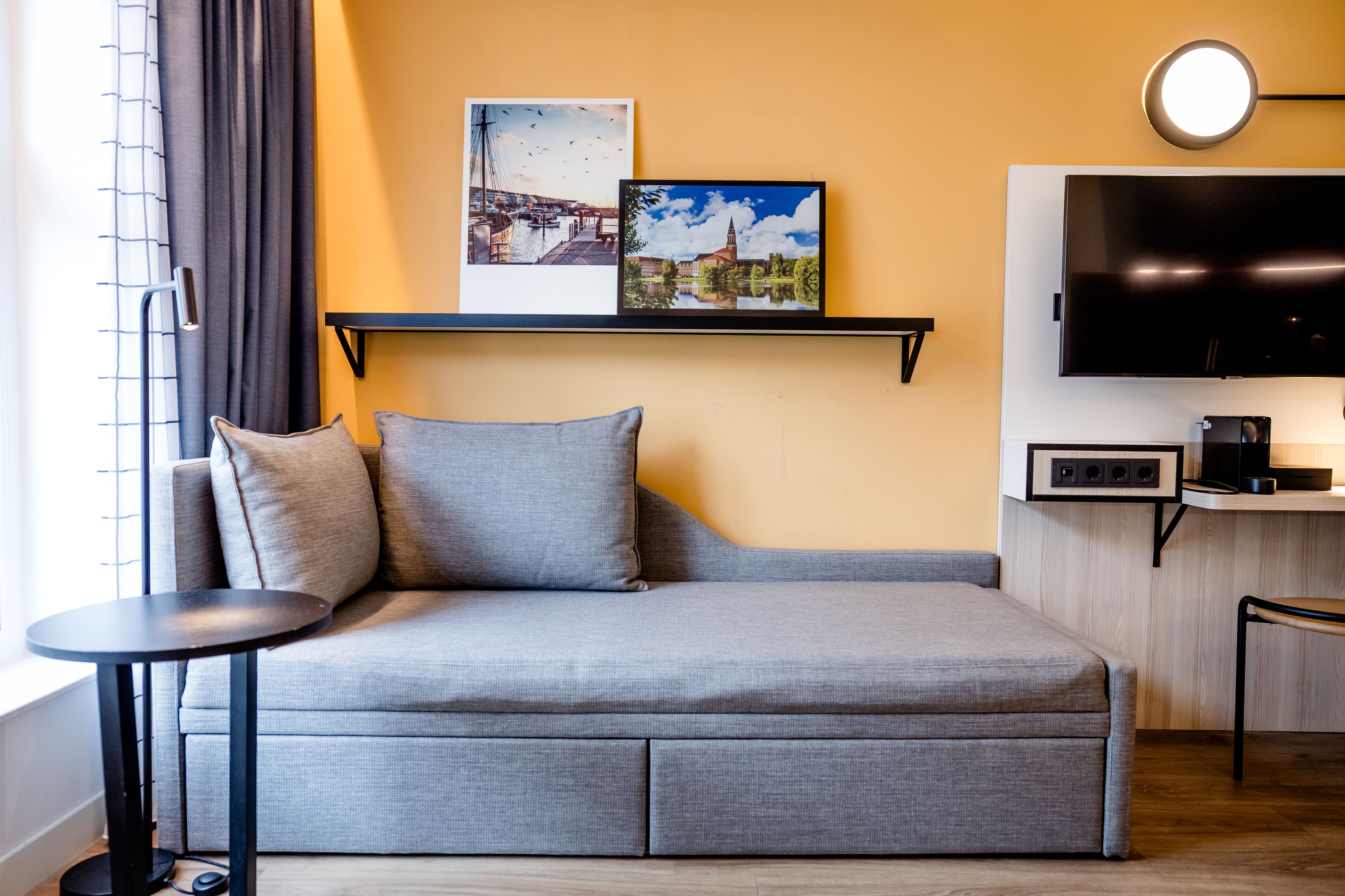 Gemütliche Sitzecke mit grauem Sofa, zwei Kissen und kleinem Beistelltisch vor sonnengelber Wand, dekoriert mit zwei Bildern. Nebenan moderner Bereich mit Wandfernseher und Arbeitsplatz, helle Raumgestaltung mit natürlichem Licht von links.