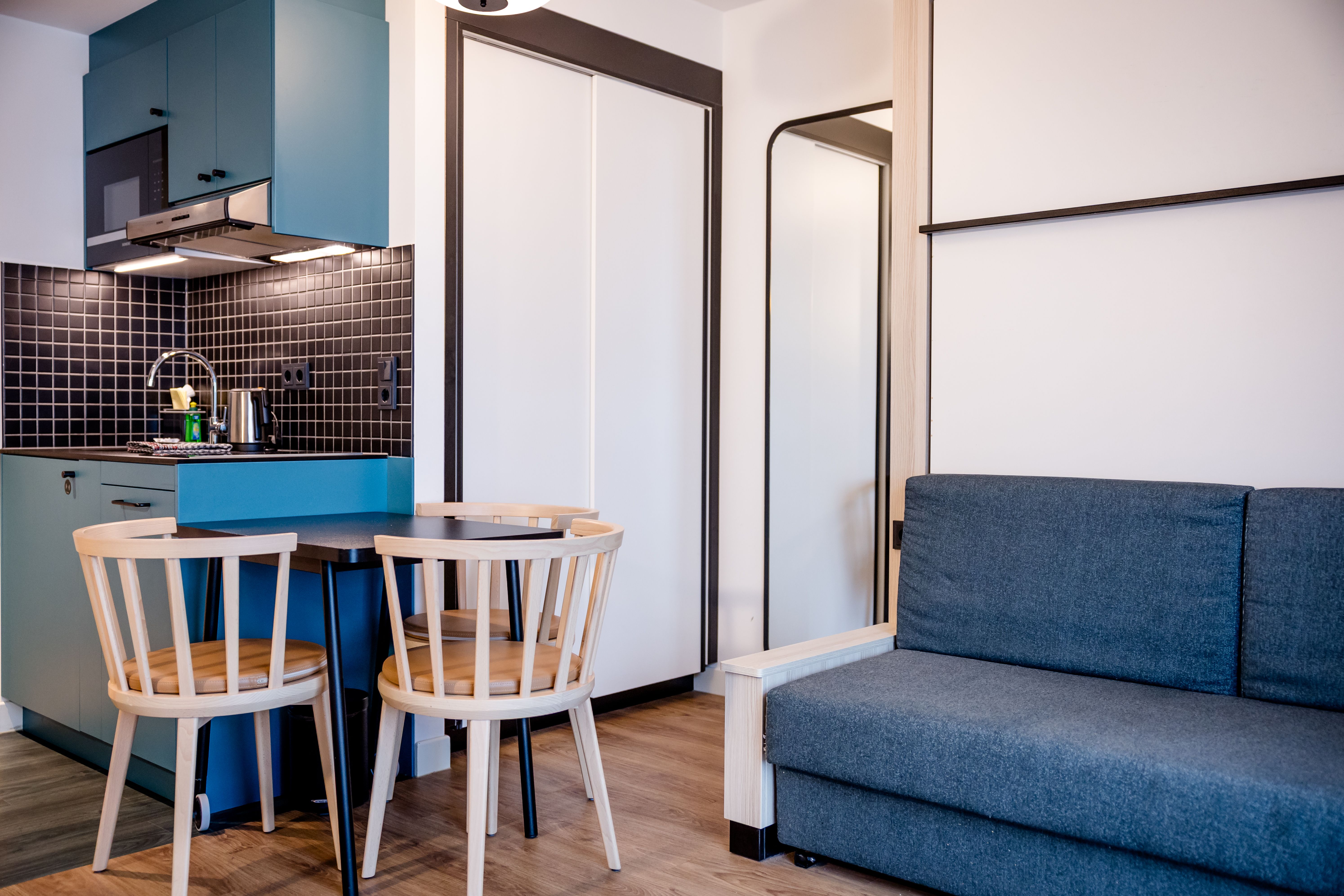 Modern eingerichtetes Apartment mit kleiner Küchenzeile, zwei Holzstühlen am Esstisch und einem blauen Sofa, das Gemütlichkeit und funktionalen Komfort bietet.