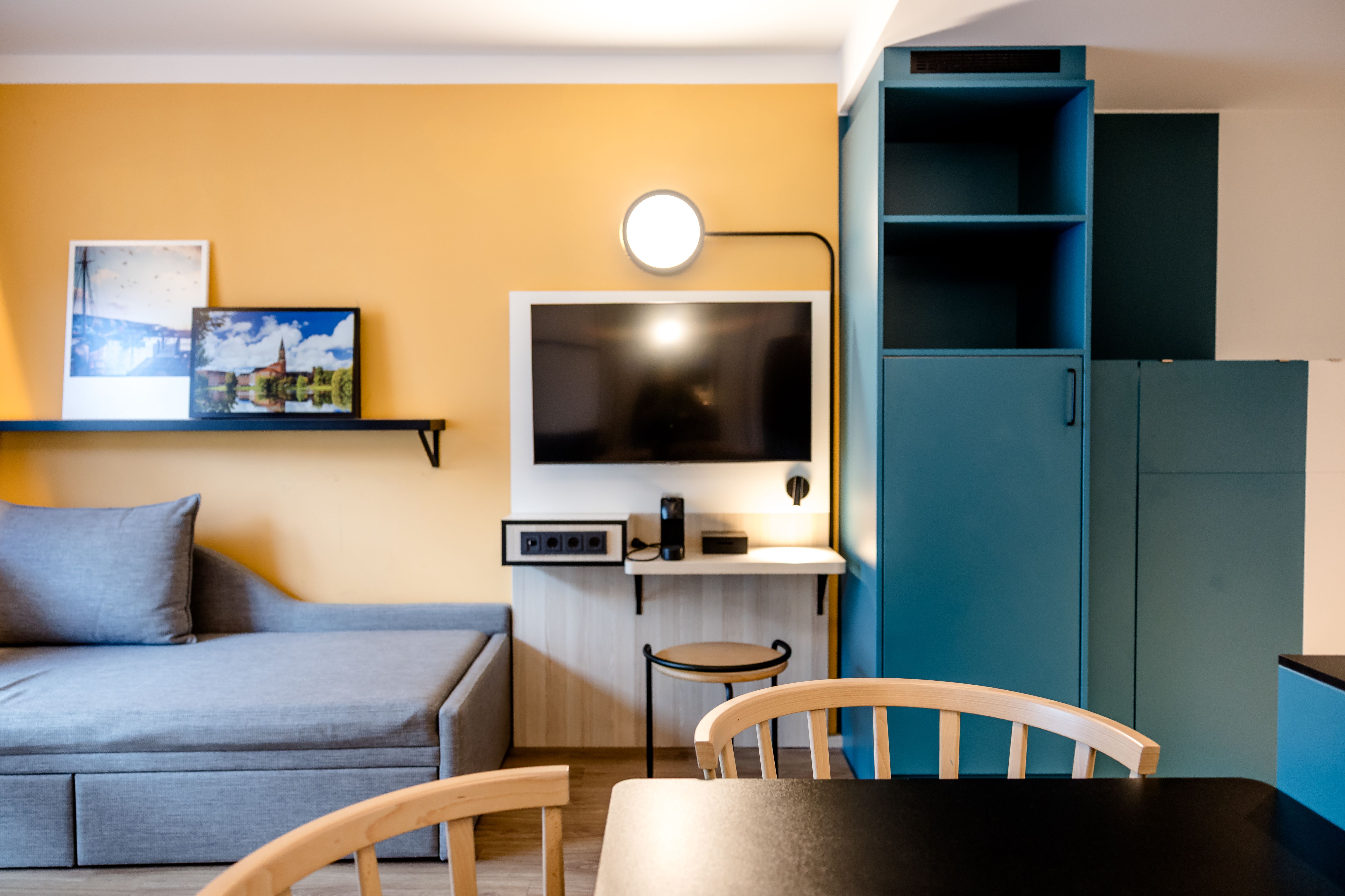 Modern eingerichtetes Hotelzimmer mit grauem Sofa, Wand-TV, Schreibtisch, Stuhl und blau-grünen Schränken. Helle Wandfarben und gemütliche Atmosphäre laden zum Entspannen ein.