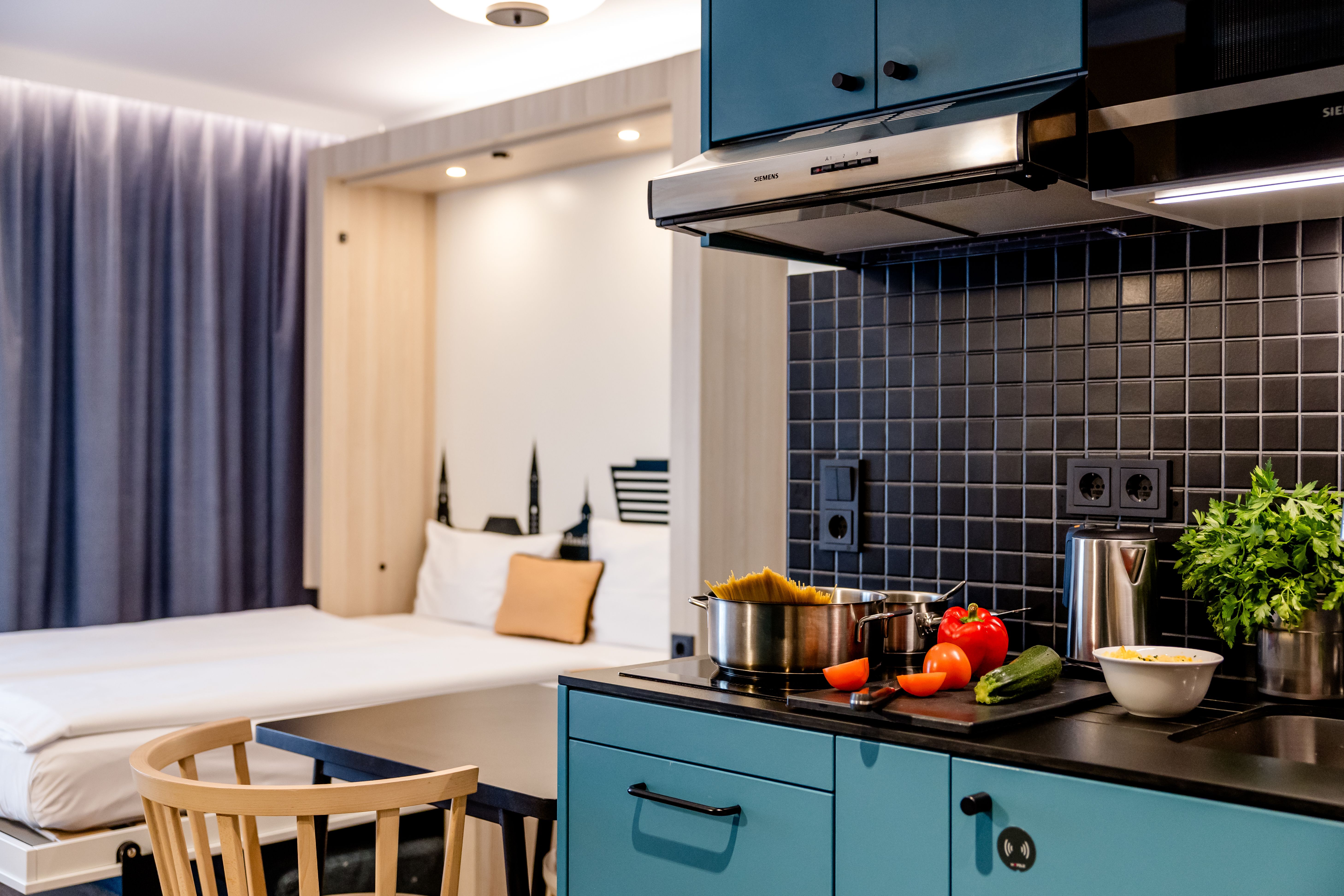Moderne Hotelzimmer-Studio mit offener Küche, Kochutensilien und frischem Gemüse auf der Arbeitsplatte, gemütlichem Doppelbett im Hintergrund, angenehmer Beleuchtung und Sitzgelegenheit, ideal für längere Aufenthalte mit Selbstverpflegung.