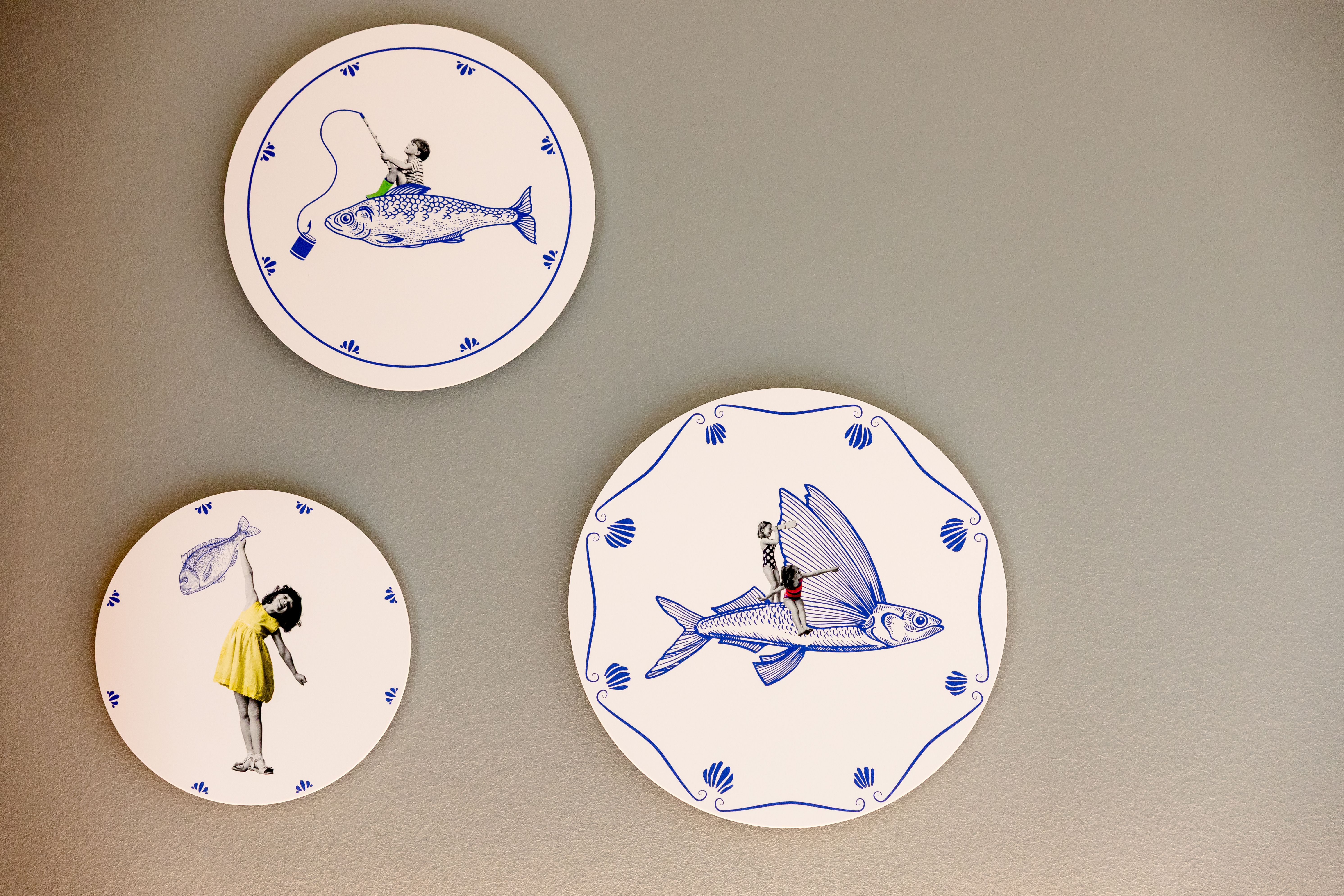 Drei dekorative Teller an einer Wand zeigen jeweils eine kleine Figur, die mit überdimensionalen Fischen interagiert – spielerisch, fantasievoll und künstlerisch gestaltet.