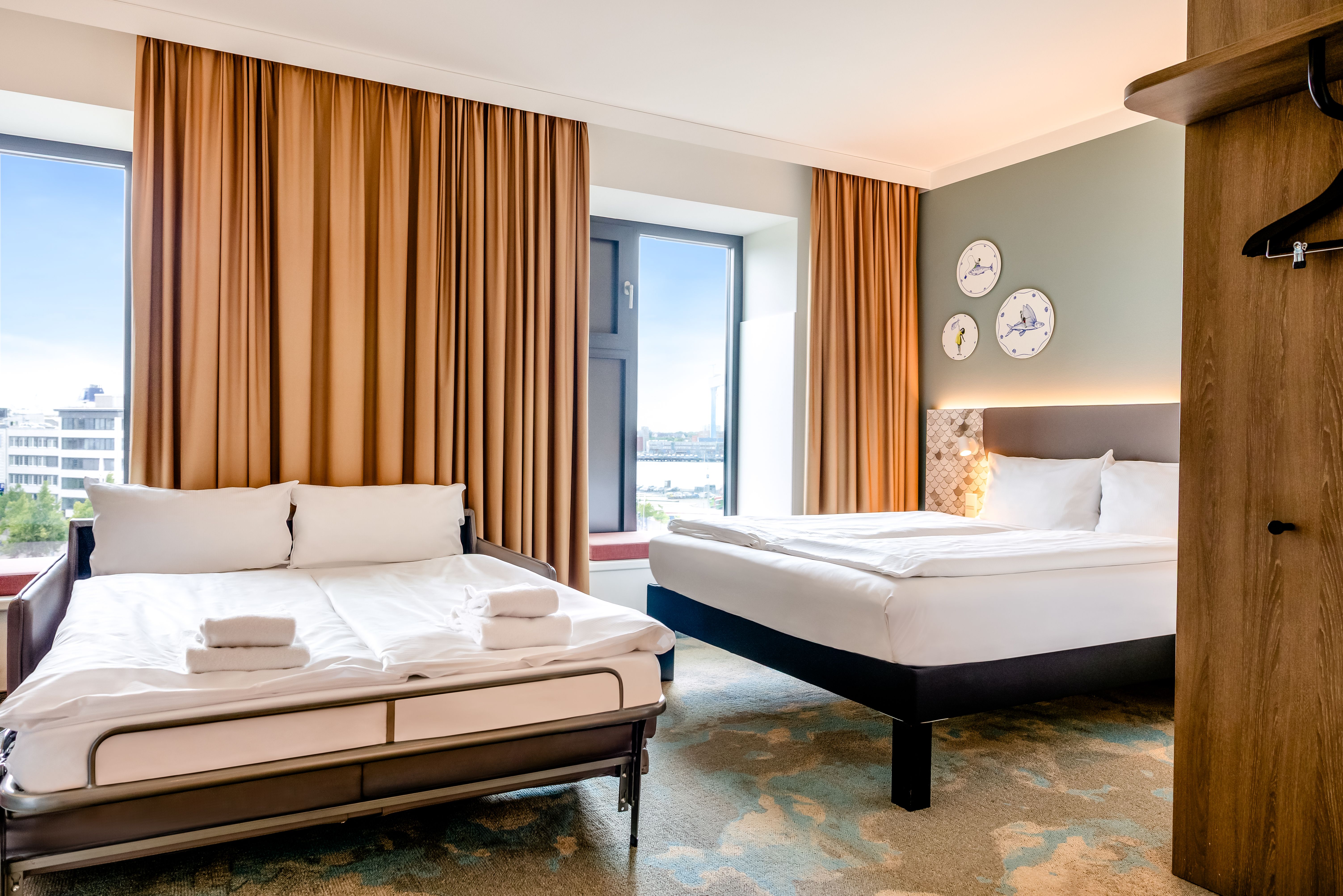 Hell eingerichtetes Hotelzimmer mit zwei großen Betten, warmen Vorhängen und Fenstern mit Blick auf die Stadt, beruhigendem Farbschema und modernen, komfortablen Möbeln für entspannte Übernachtungen.