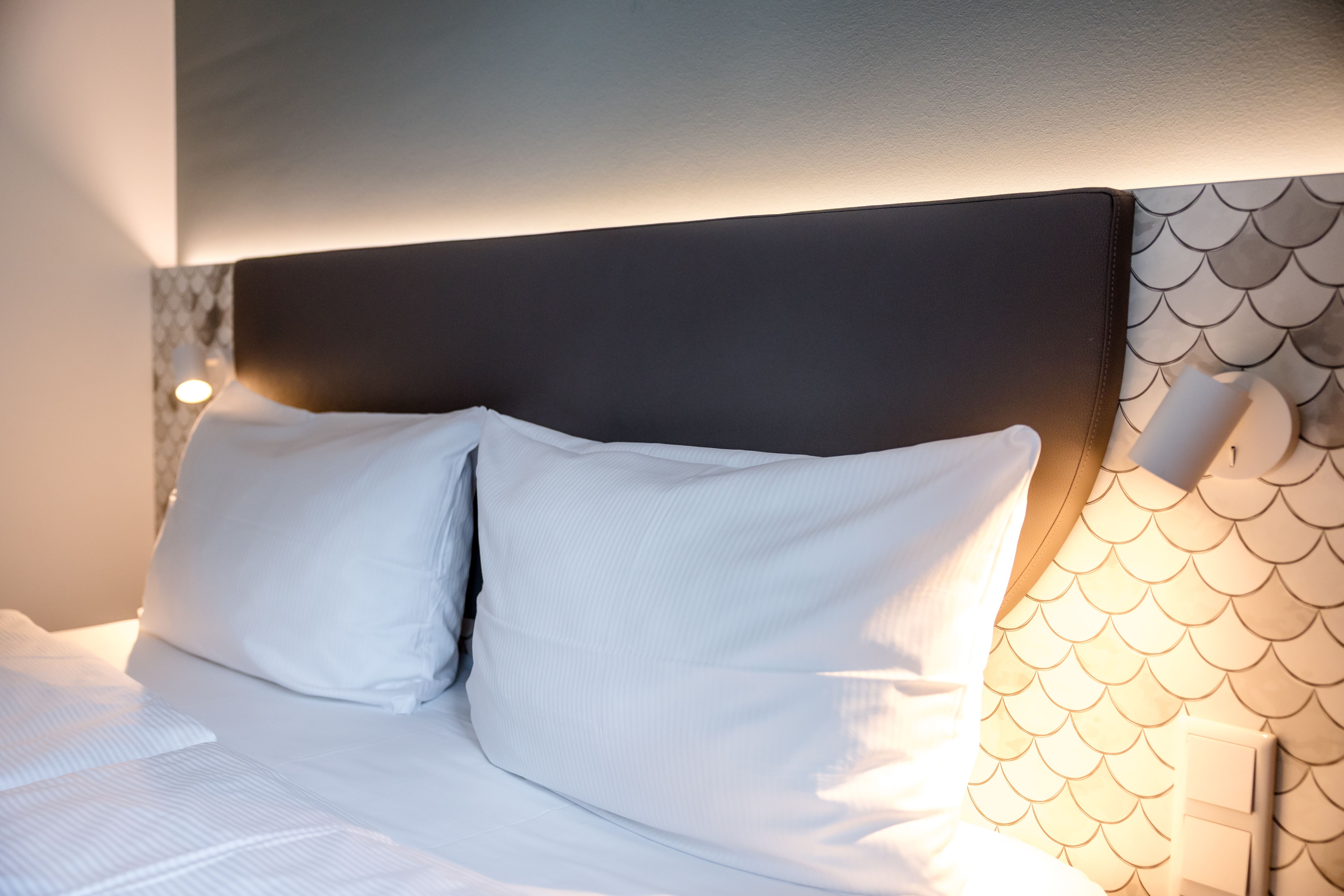 Bequemes Doppelbett mit weißen Kissen und Bettwäsche, gedämpfte Beleuchtung mit indirektem Licht und modernem Wanddesign, das eine entspannte und einladende Atmosphäre im Zimmer schafft.