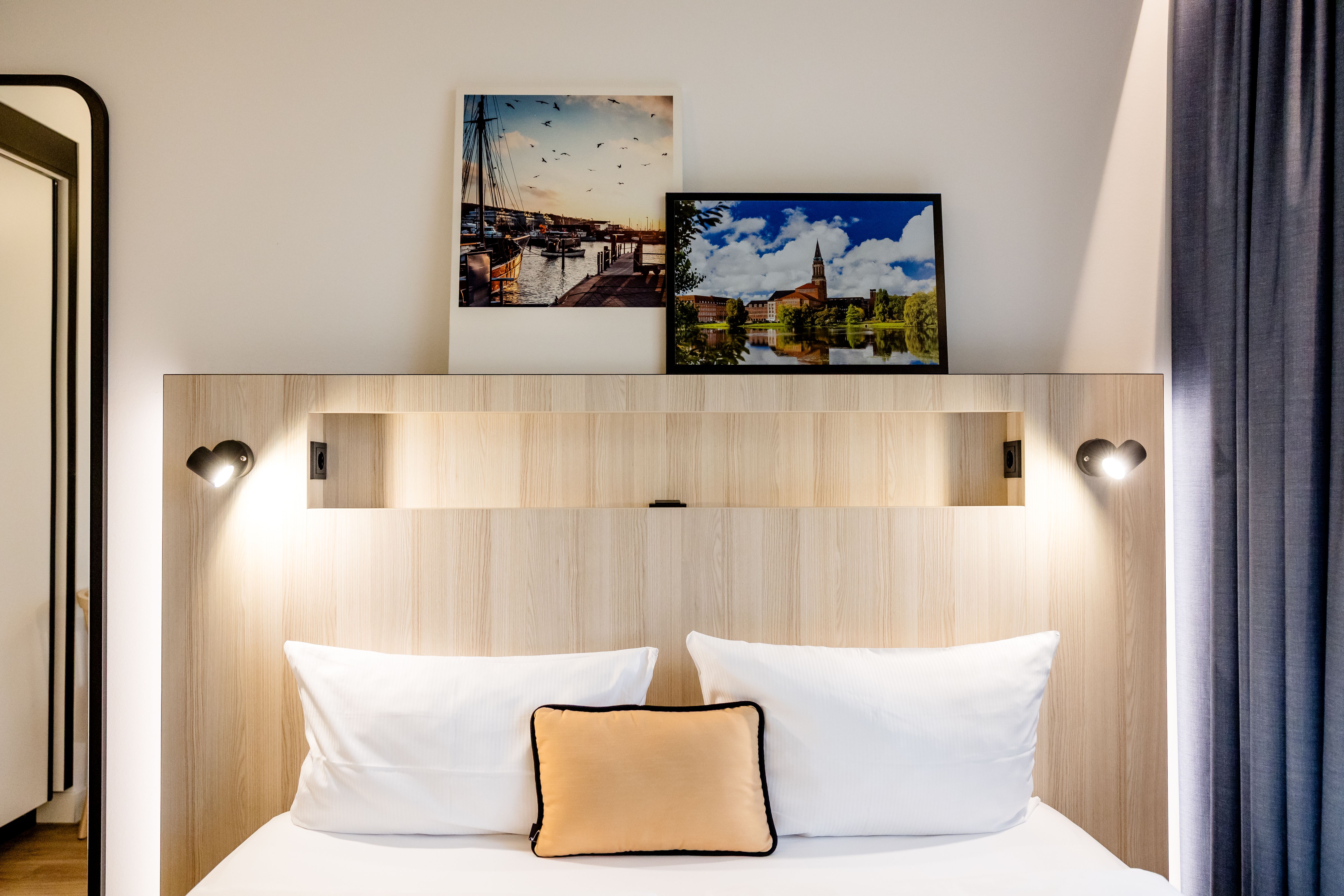 Modern eingerichtetes Hotelzimmer mit Doppelbett, weißen Kissen und Akzentkissen, dezenter Beleuchtung und stimmungsvollen Landschaftsbildern an der Wand, die eine ruhige und einladende Atmosphäre schaffen.