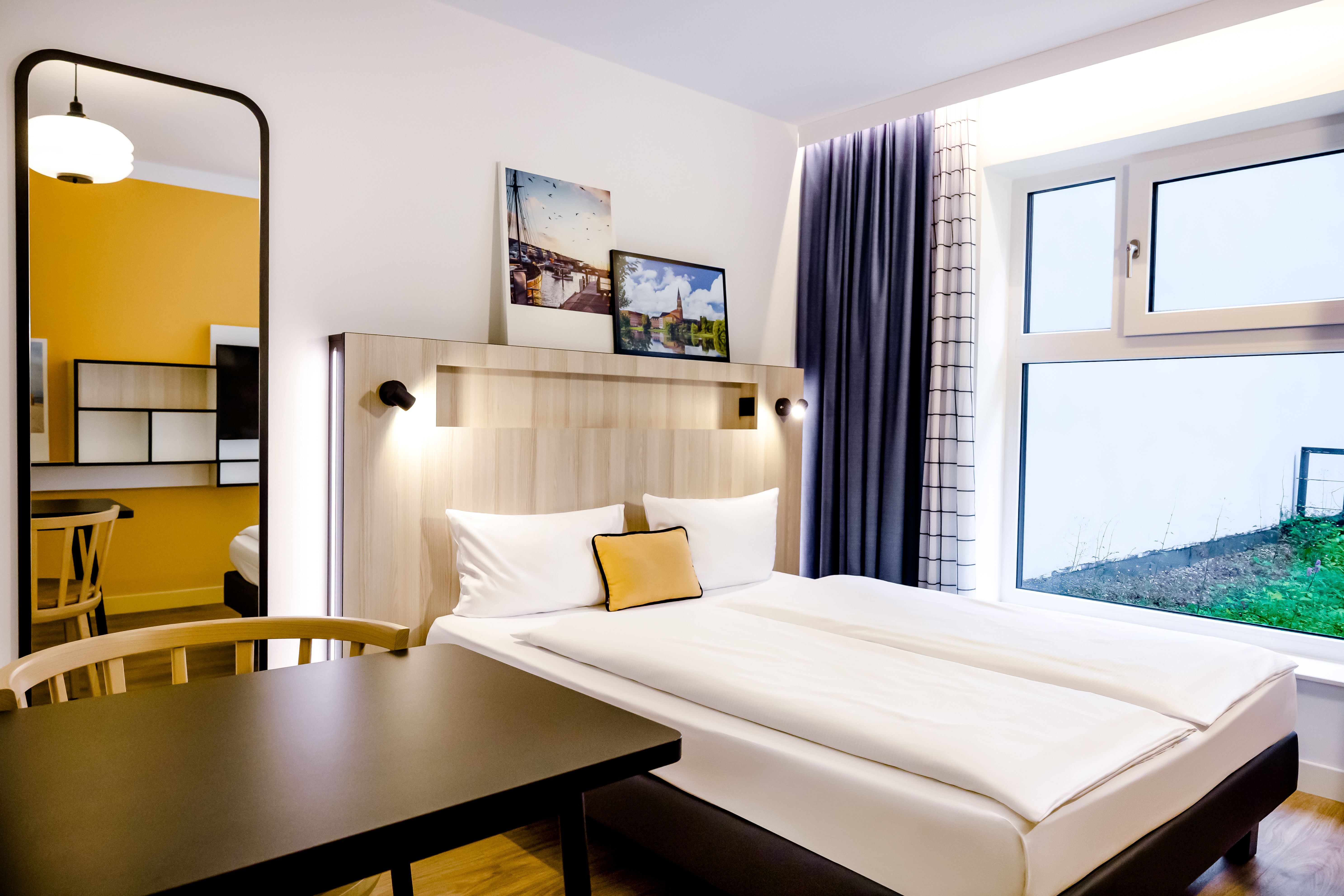 Helles, modern eingerichtetes Hotelzimmer mit großem Fenster, gemütlichem Doppelbett, stilvollem Holzmobiliar und warmen Farbakzenten, ideal für erholsamen Aufenthalt.