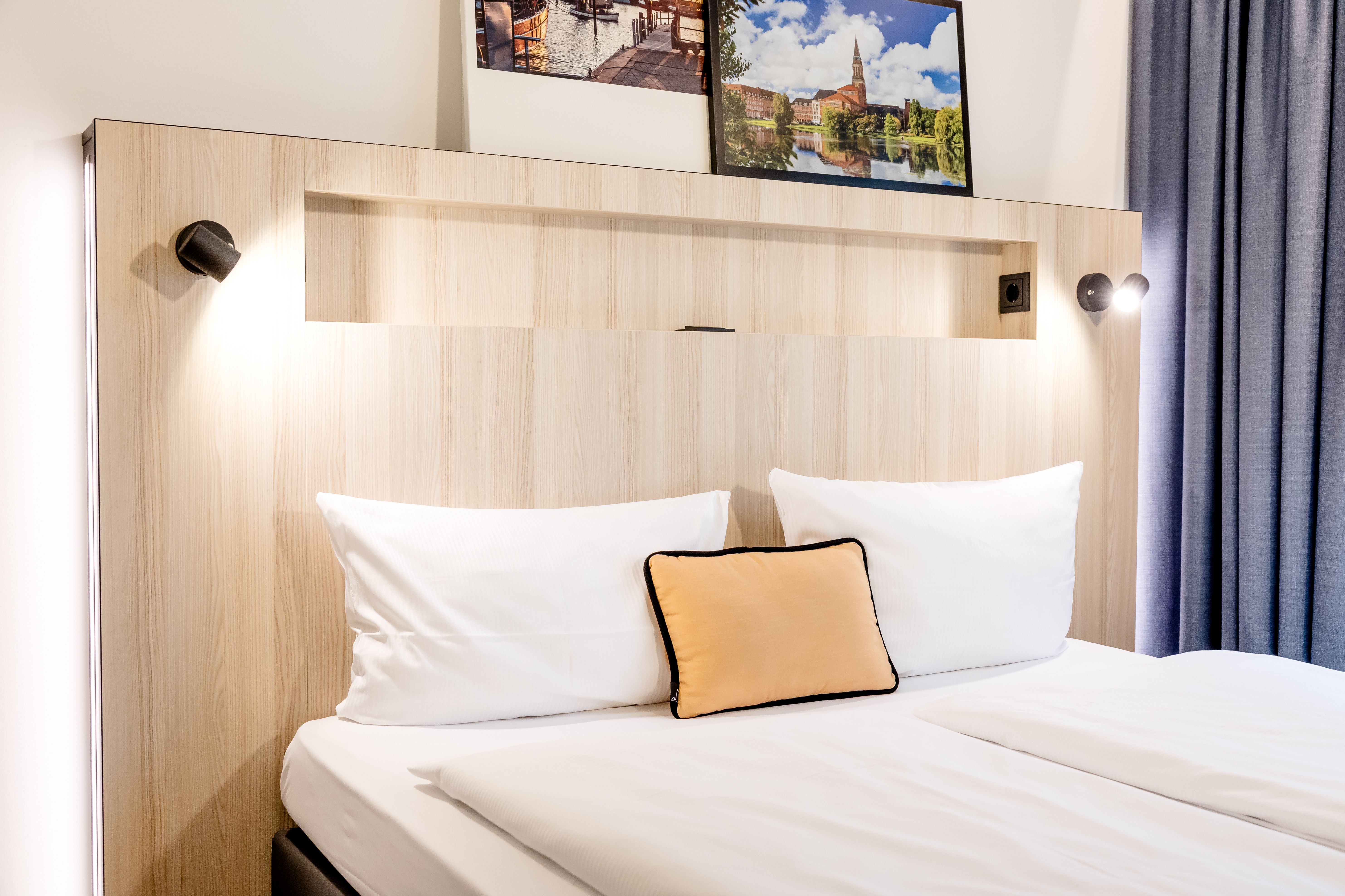 Modern eingerichtetes Hotelzimmer mit hellem Holzkopfteil, zwei weißen Kissen, einem orangefarbenen Zierkissen und stimmungsvoller Beleuchtung für entspannte Nächte.