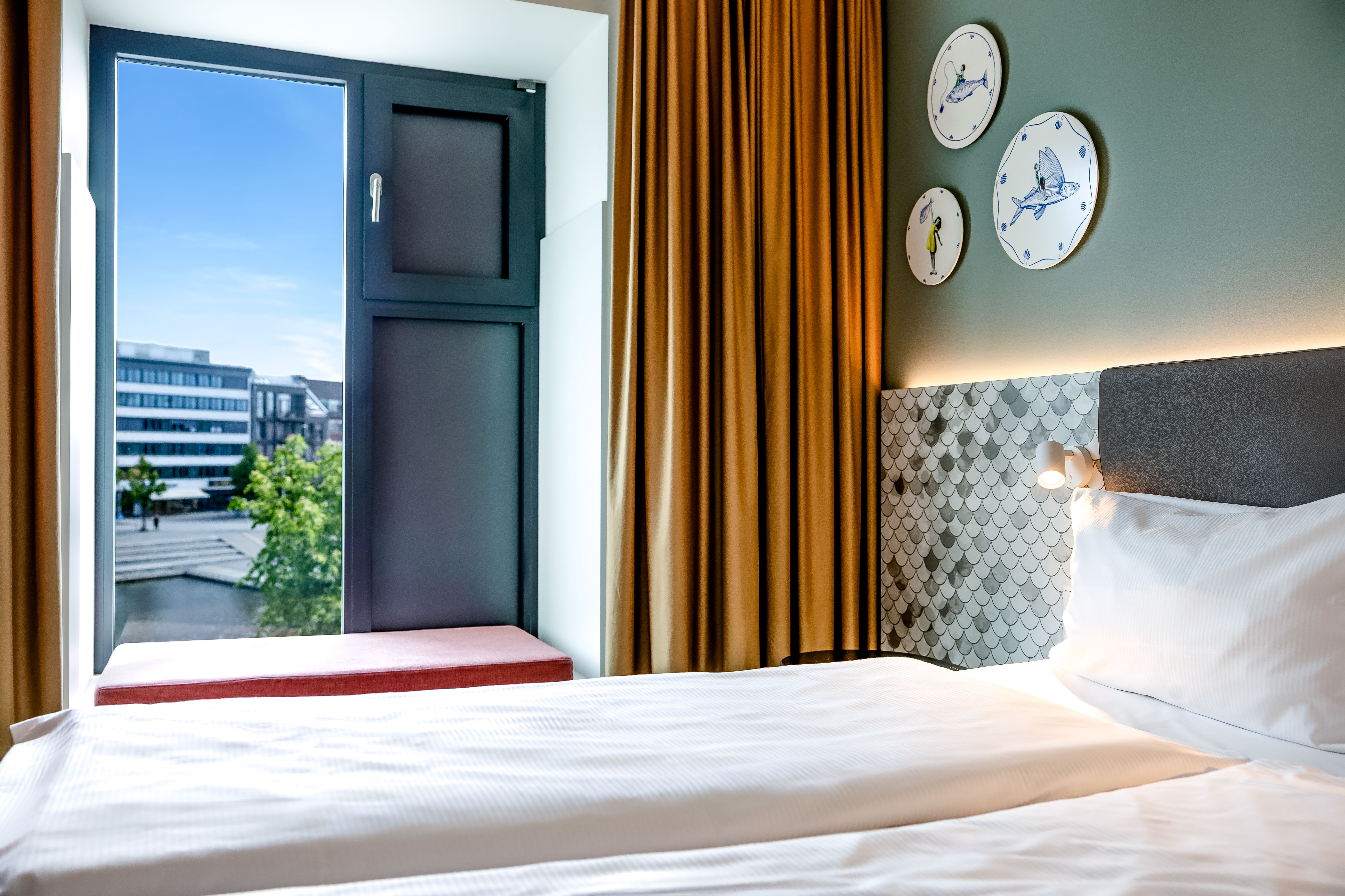 Helles Hotelzimmer mit großem Fenster, ruhigem Stadtblick, gemütlichem Bett und warmen Vorhängen, das eine entspannte Atmosphäre für erholsamen Schlaf und Tageslicht bietet.