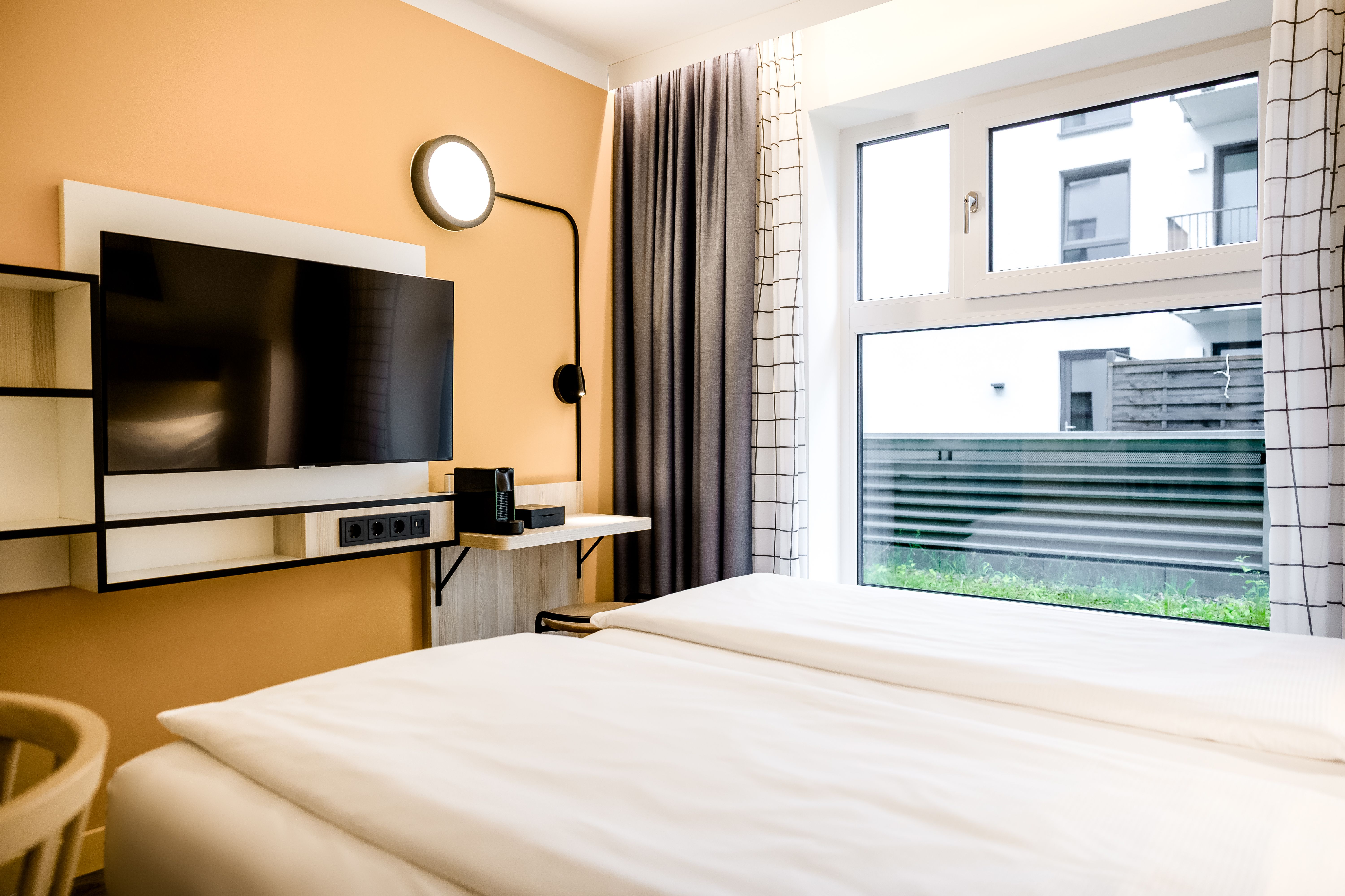 Helles Hotelzimmer mit gemütlichem Doppelbett, großem Fenster für Tageslicht, modernem Flachbildfernseher an orangefarbener Wand und klaren, hellen Vorhängen, das eine einladende und komfortable Atmosphäre schafft.