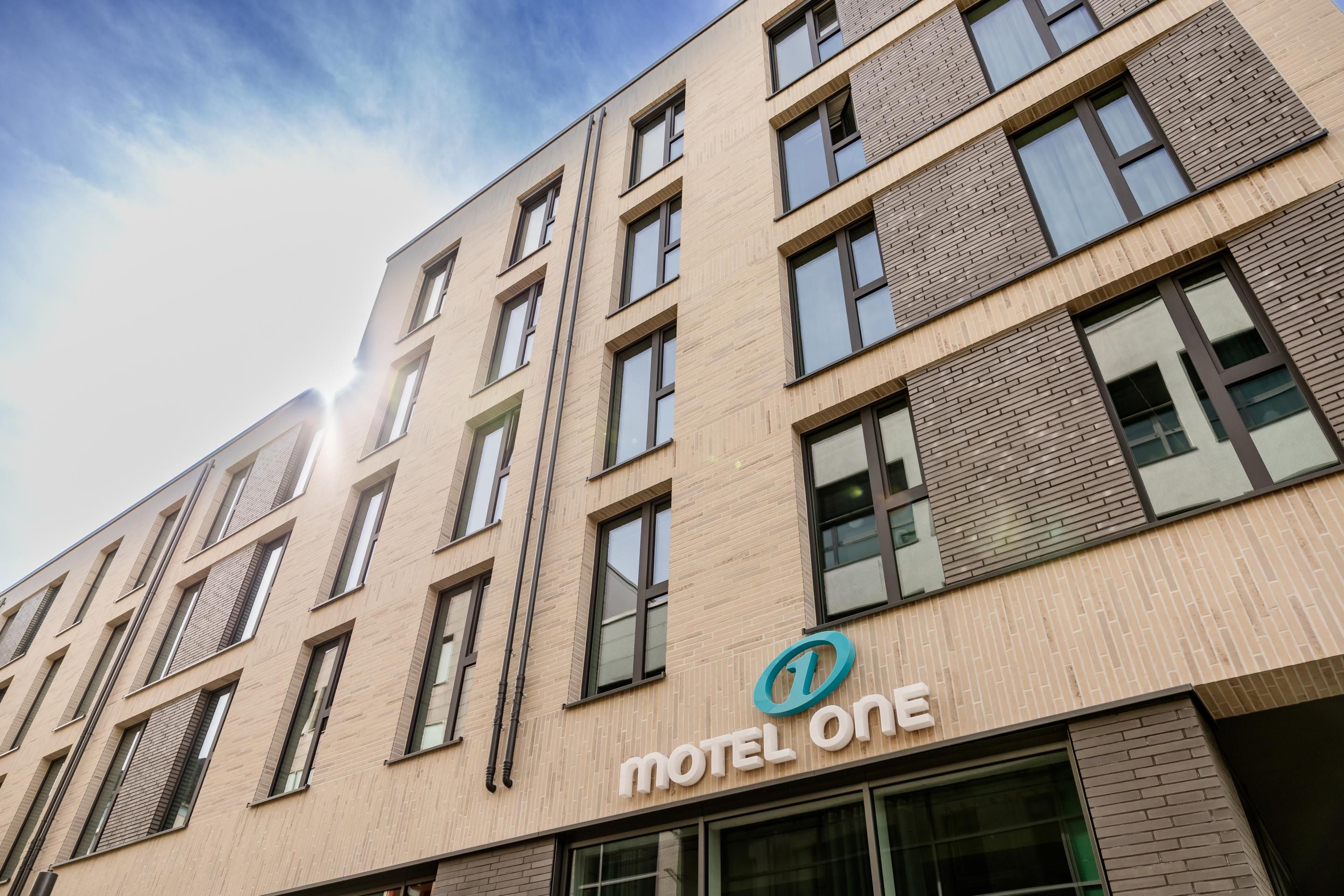 Fassade eines modernen Motel One Gebäudes mit großen Fenstern und hellem, kombiniertem Stein- und Ziegelfrontdesign, aufgenommen bei Sonnenlicht.
