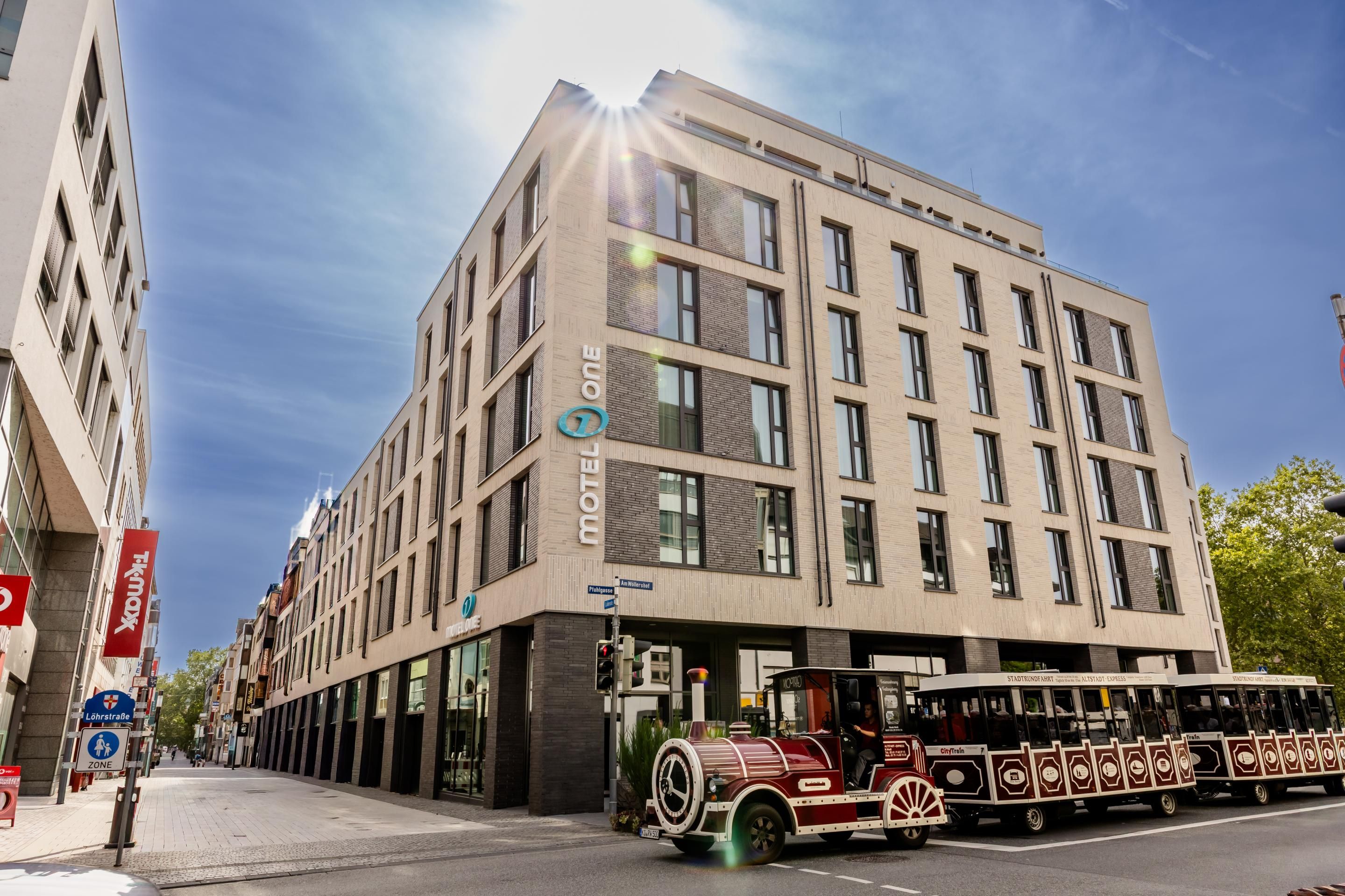 Modernes, mehrstöckiges Hotelgebäude unter blauem Himmel an einer Straßenecke, vor dem eine kleine touristische Straßenbahn hält.