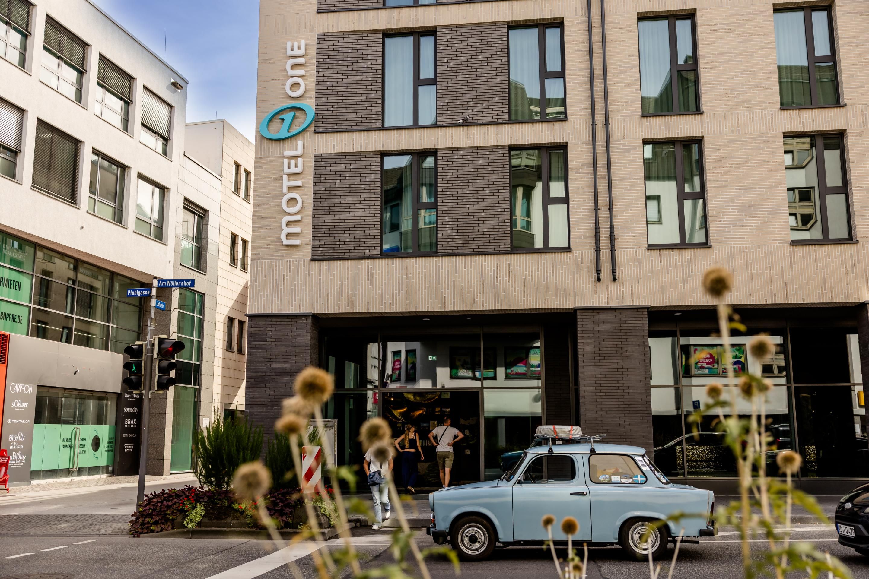 Außenansicht des modernen Motel One mit klarer Fassade und großen Fenstern, im Vordergrund ein historisches Auto und städtisches Straßenbild.