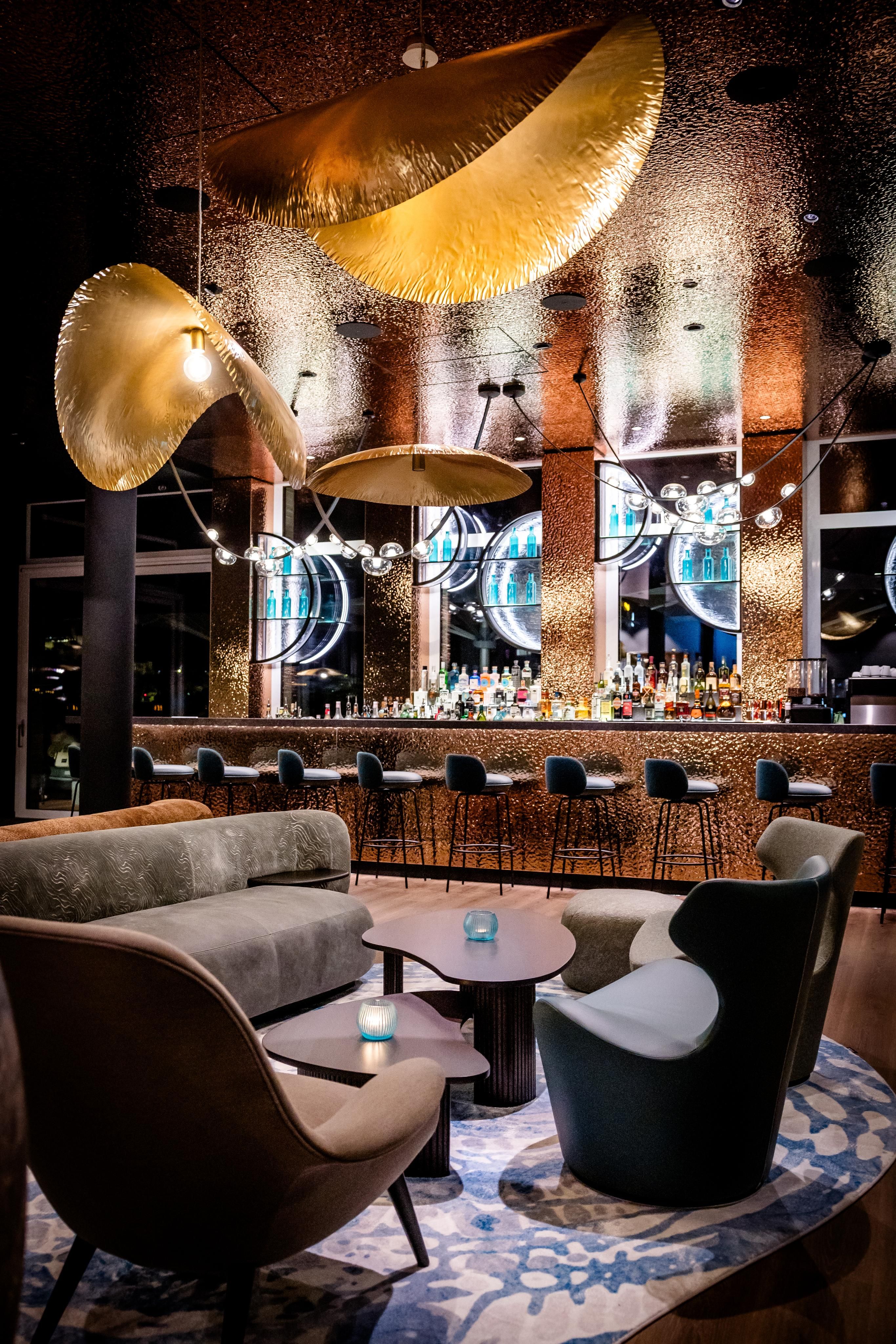 Moderne Hotelbar mit stilvollen Sitzgruppen, warmen goldfarbenen Deckenelementen und einer gut bestückten Bar, die durch indirekte Beleuchtung eine einladende Atmosphäre schafft.