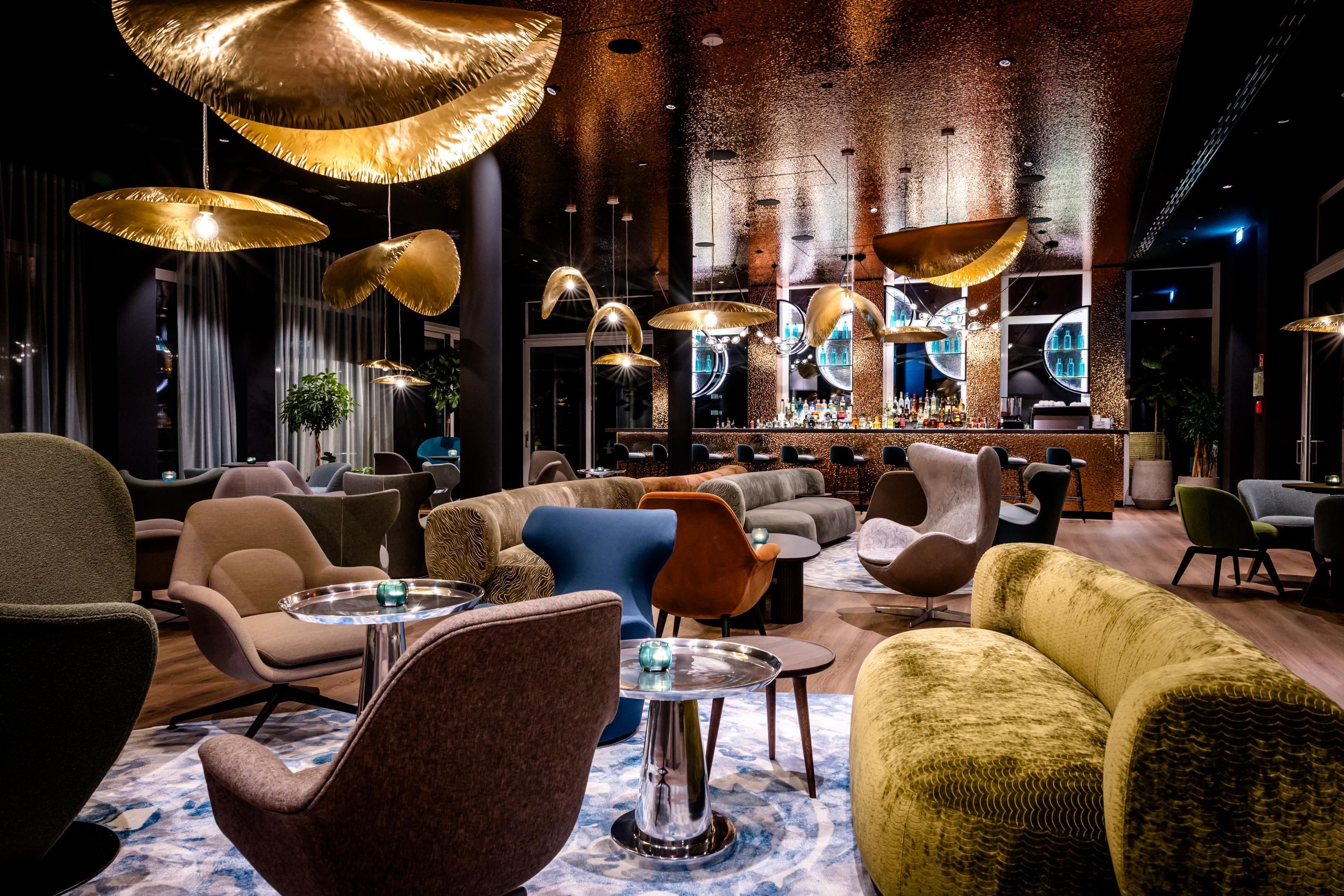Moderne Lounge mit stilvollen, bequemen Sesseln und Sofas, warmem Licht durch goldene, futuristische Deckenlampen, ruhiger Atmosphäre und einladendem Ambiente für entspannte Abende oder lockere Gespräche.