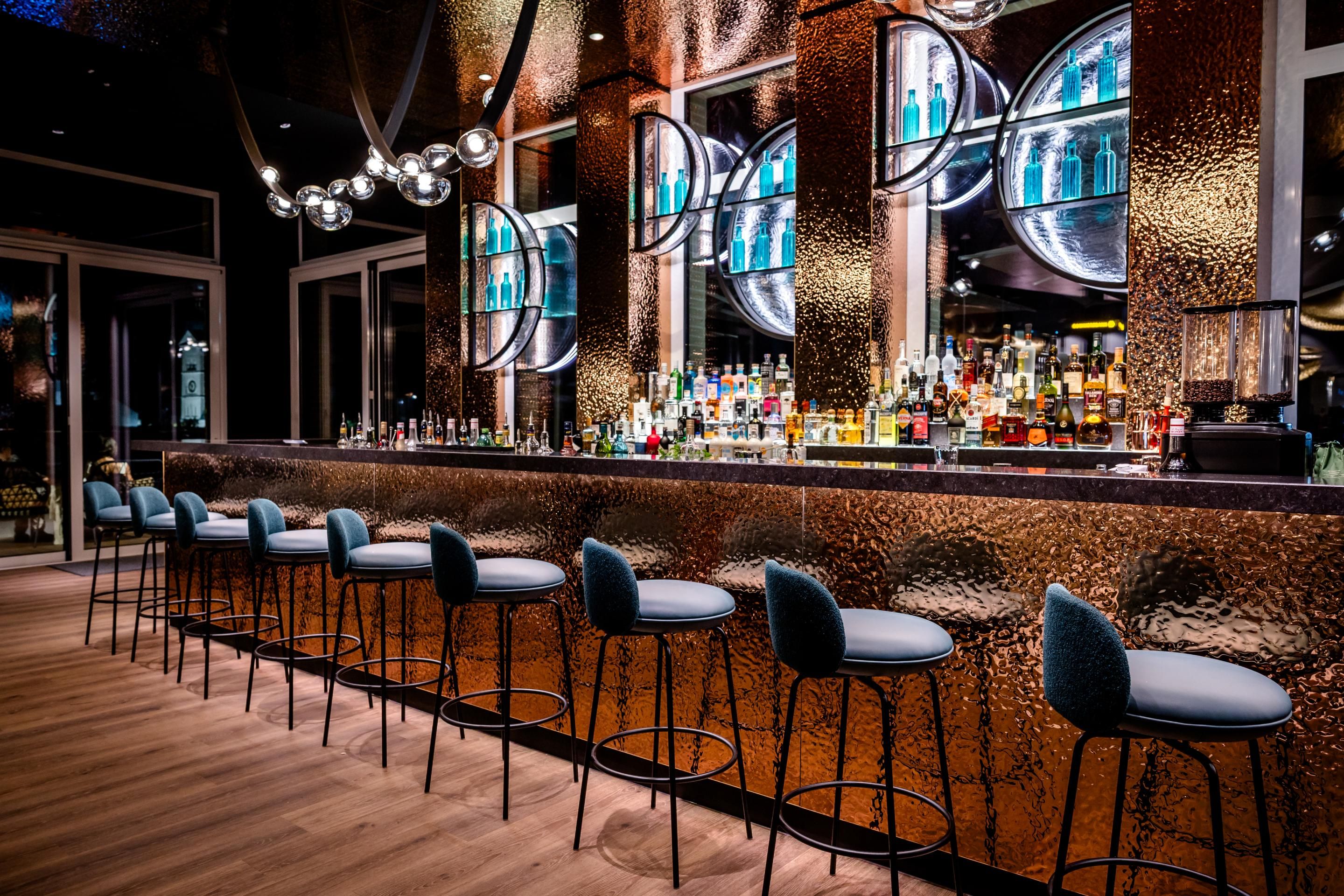 Elegante Hotelbar mit langem Tresen, acht bequemen Barhockern, stimmungsvoller Beleuchtung und stilvollen Spiegeln. Modernes Ambiente für entspannte Abende und gesellige Treffen.
