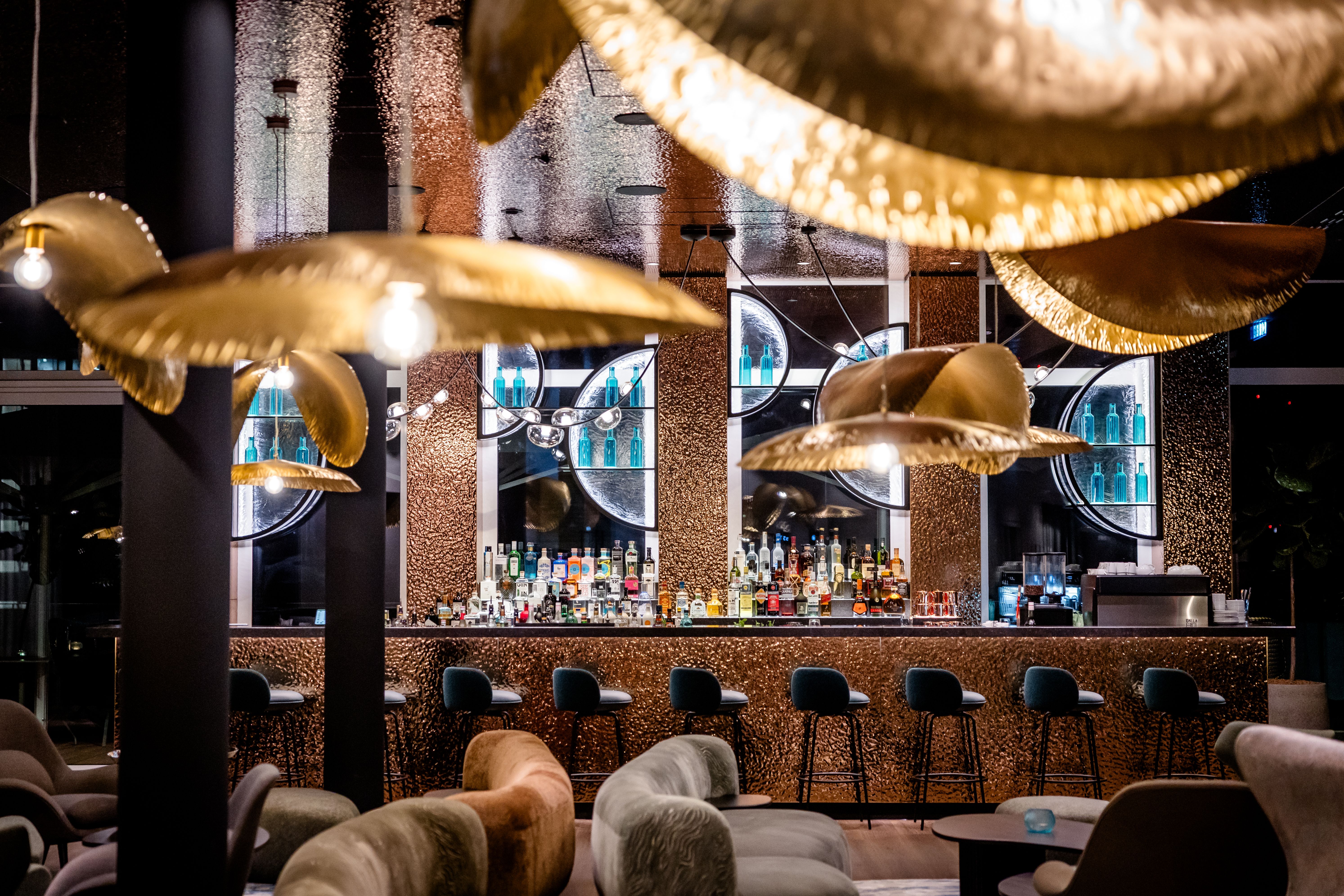 Moderne Hotelbar mit gemütlichen Sitzbereichen, hohen Barhockern und stilvollen, goldfarbenen Hängelampen, die eine elegante und einladende Atmosphäre schaffen. Spiegel und beleuchtete Regale mit Spirituosen betonen das luxuriöse Ambiente. Ideal für entspannte Abende und gesellige Treffen.