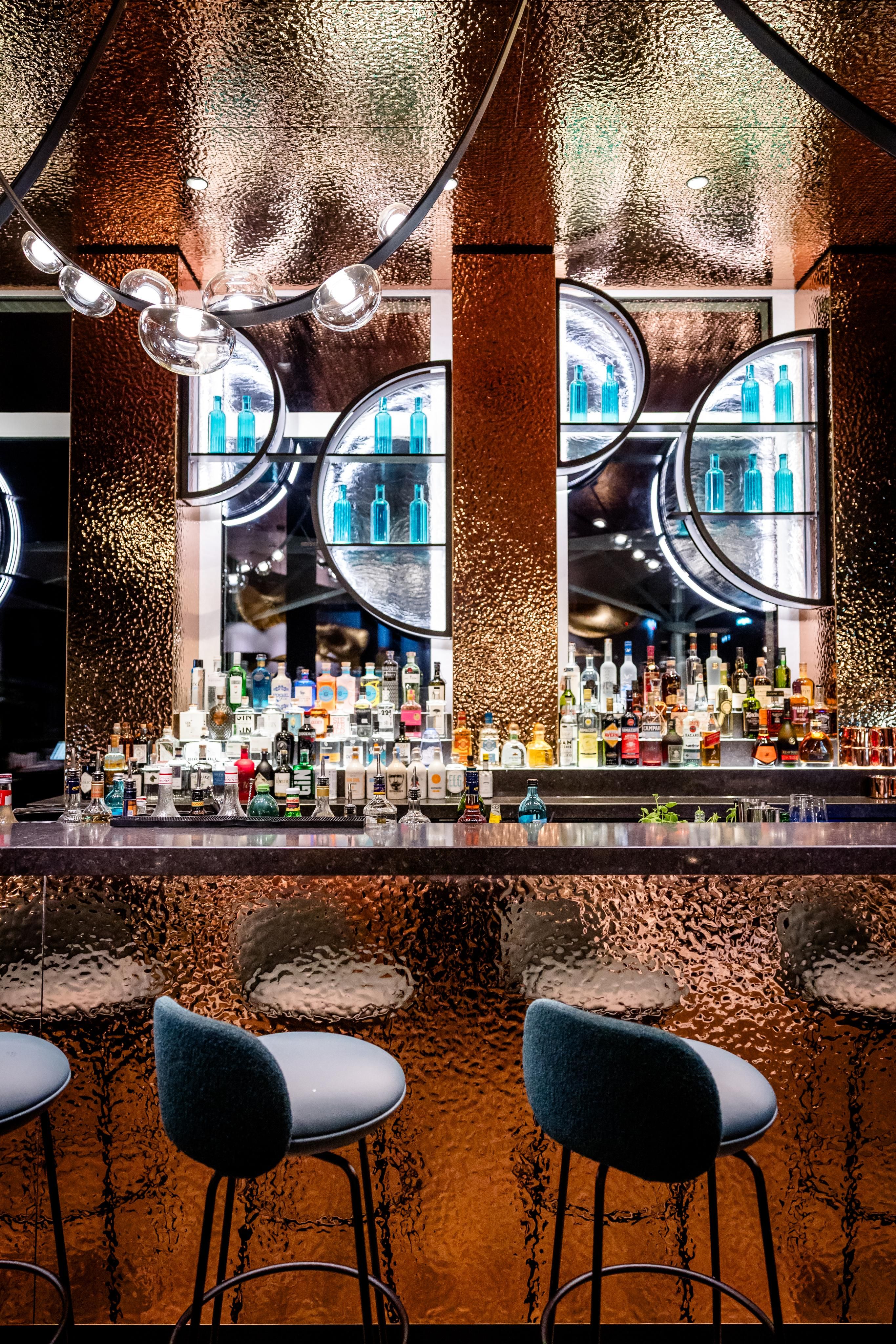 Elegante Hotelbar mit beleuchtetem Tresen, zahlreichen Spirituosen und stilvollen Barhockern, umgeben von warm reflektierenden, texturierten Wänden und modernen dekorativen Spiegeln.