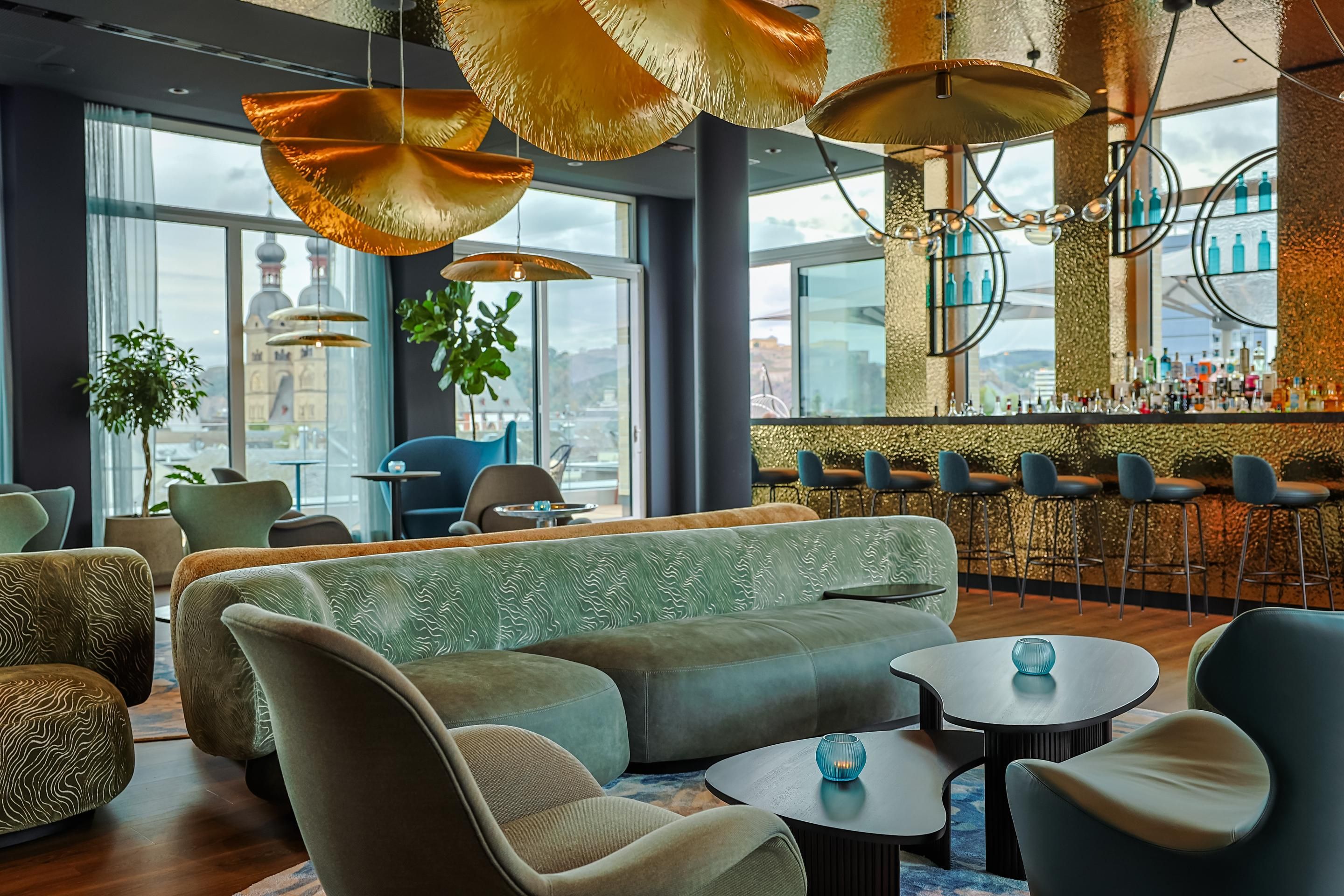 Modern gestaltete Hotelbar mit bequemen Sesseln und Sofas, großen Fenstern mit Stadtblick, auffälligen goldfarbenen Deckenleuchten und einer langen Bar mit Barhockern, die zum entspannten Verweilen einlädt.