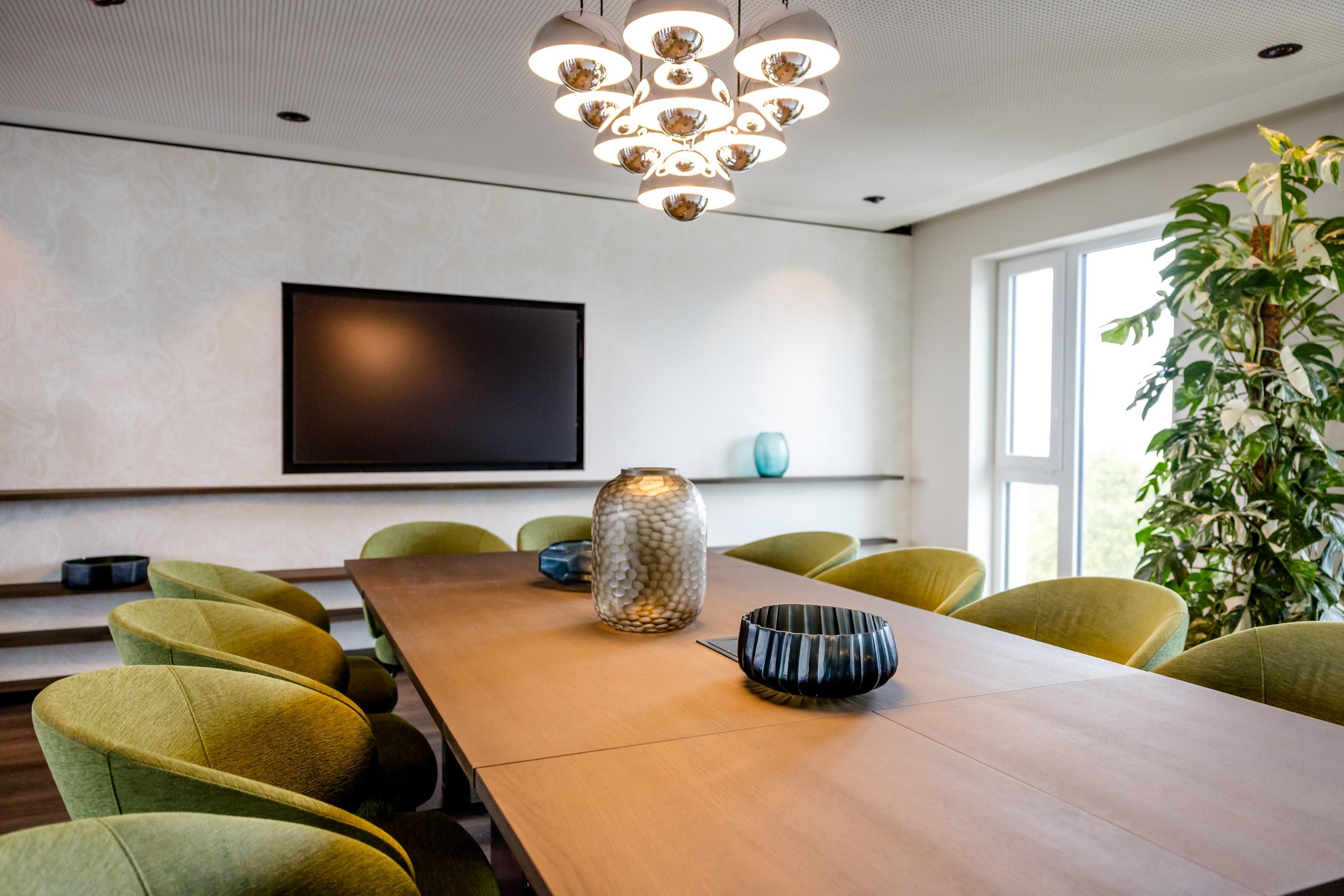 Moderner, heller Besprechungsraum mit großem Holztisch, grünen Polstersesseln, stilvoller Deckenbeleuchtung und großem Flachbildschirm an der Wand, ideal für geschäftliche Treffen.