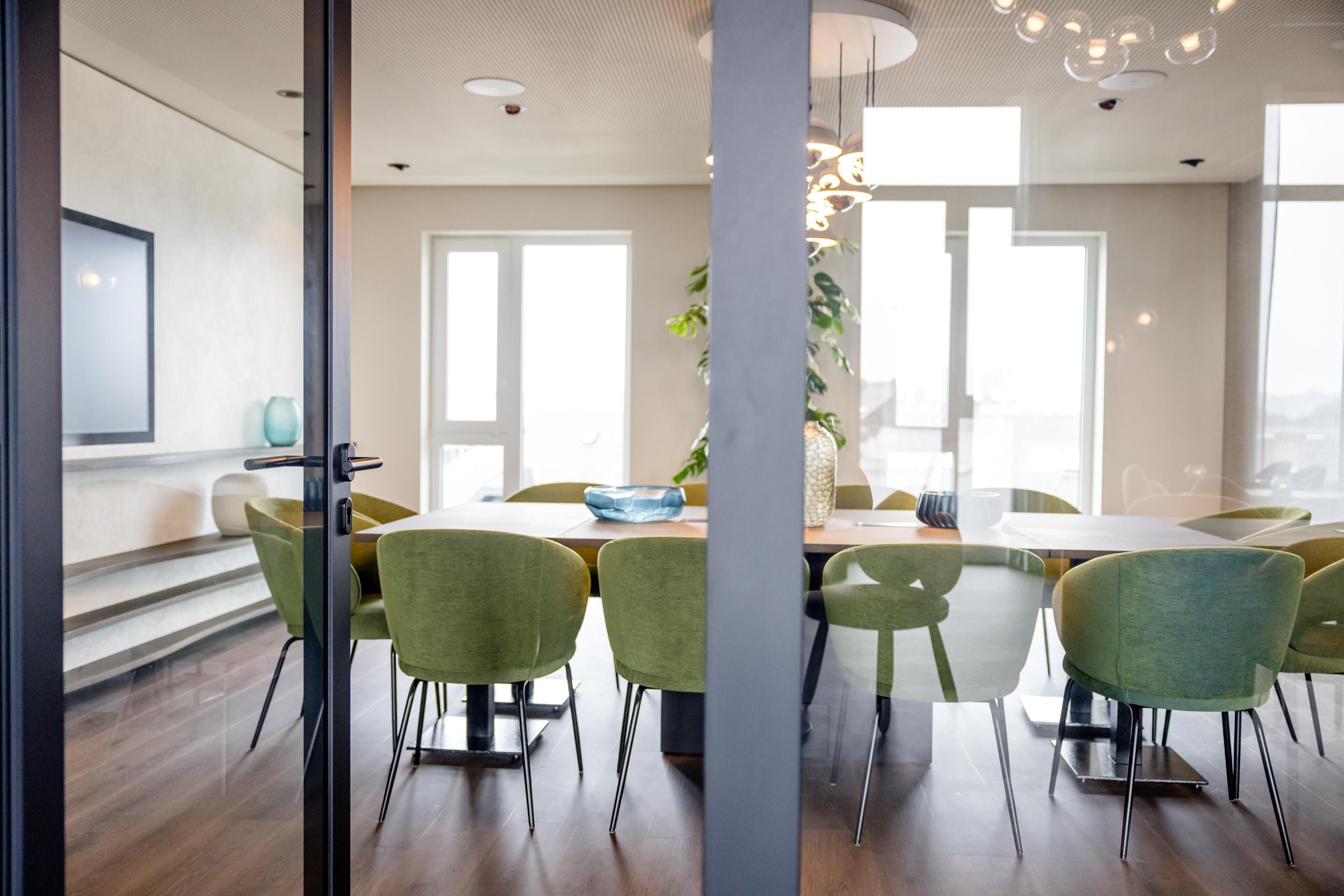Moderne, helle Tagungsraum mit großem Holztisch, umgeben von grünen Stühlen, viel Tageslicht durch große Fenster, minimalistische Dekoration, ruhige und professionelle Atmosphäre.