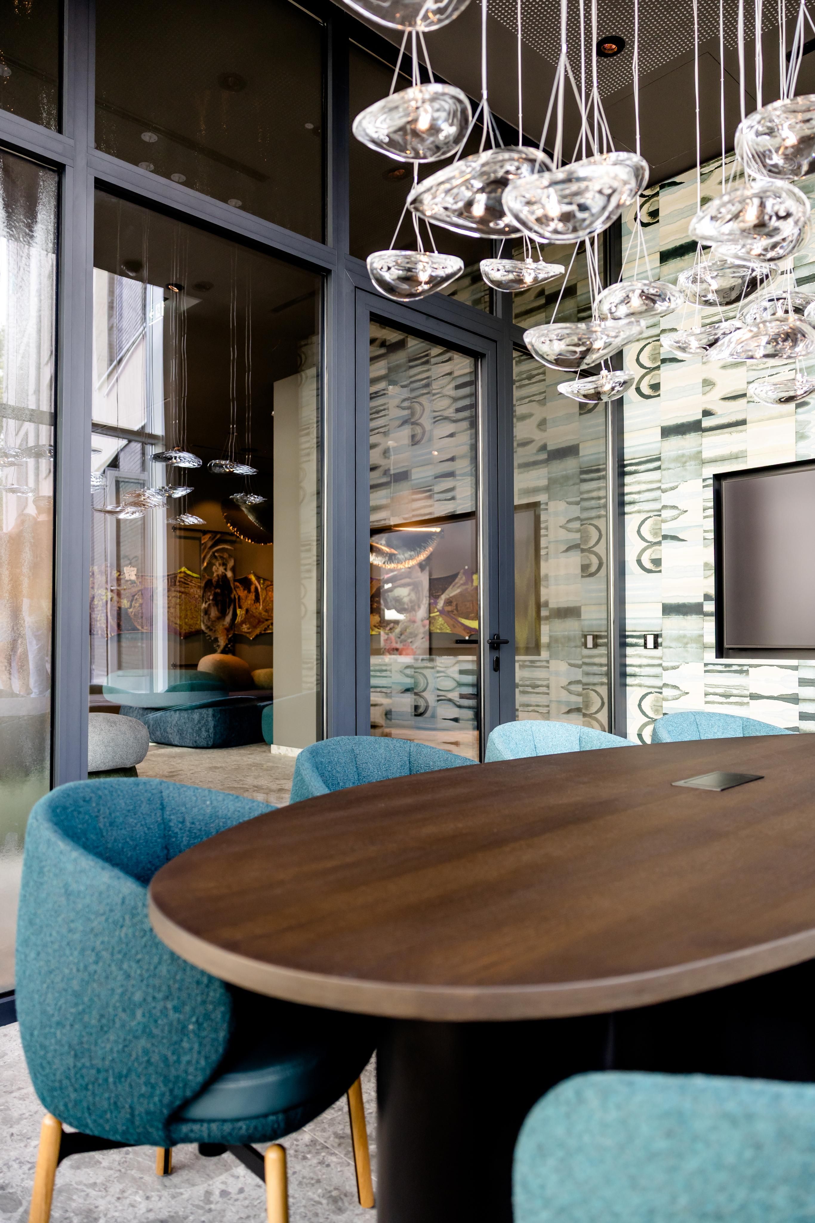 Moderner Besprechungsraum mit ovalem Holztisch, blau gepolsterten Stühlen und großem Fensterfront. Dekorative, elegante Hängeleuchte aus Glas sorgt für stimmungsvolle Beleuchtung. Raum wirkt einladend und professionell, ideal für Meetings und Gespräche.