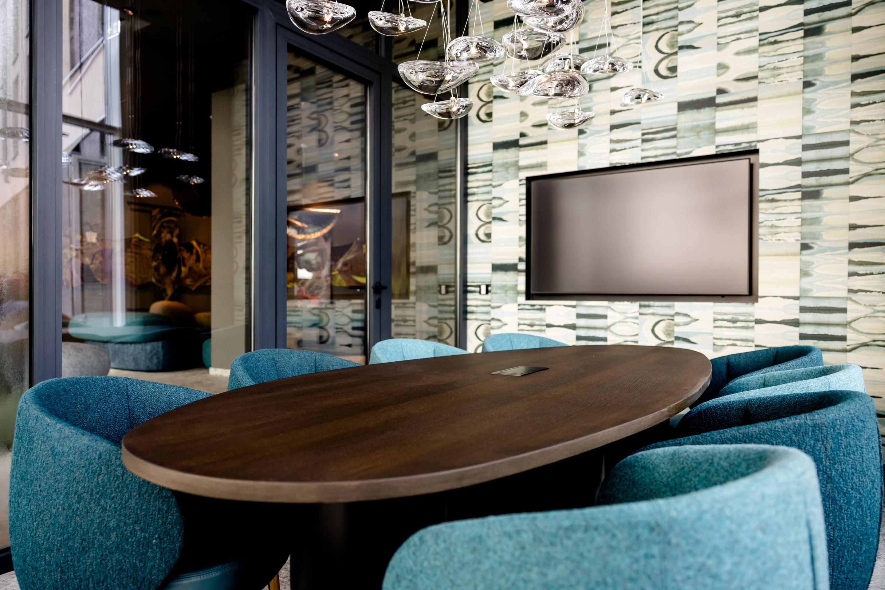 Modern eingerichteter Besprechungsraum mit ovalem Holztisch, blauen Polstersesseln und großem Wandbildschirm, umgeben von Glaswänden und dezentem, stilvollem Lichtdesign.