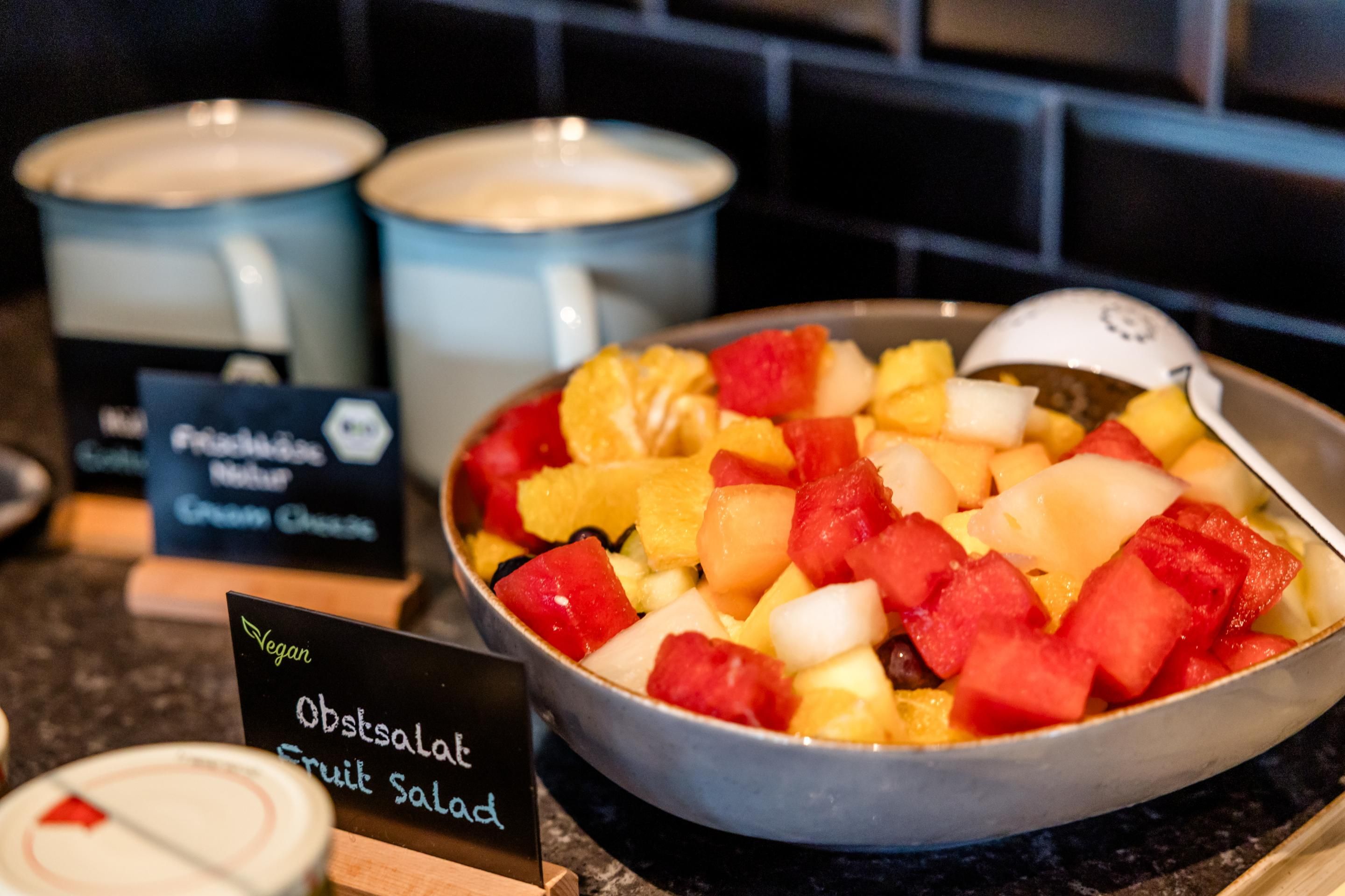 Bunte Obstsalat-Schale mit Melone, Ananas und anderen Früchten, daneben zwei Becher Hafermilch, beschriftet als vegane Frühstücksoption.