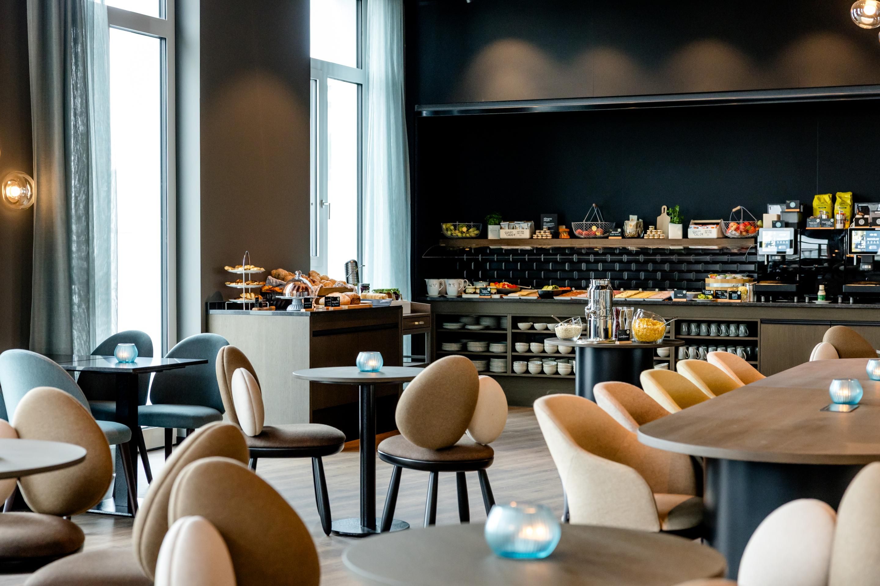 Modernes Frühstücksbuffet in hellem, stilvollem Raum mit gemütlichen Stühlen und Tischen, große Fenster für viel Tageslicht, vielfältige Auswahl an Speisen und Getränken für einen angenehmen Start in den Tag.