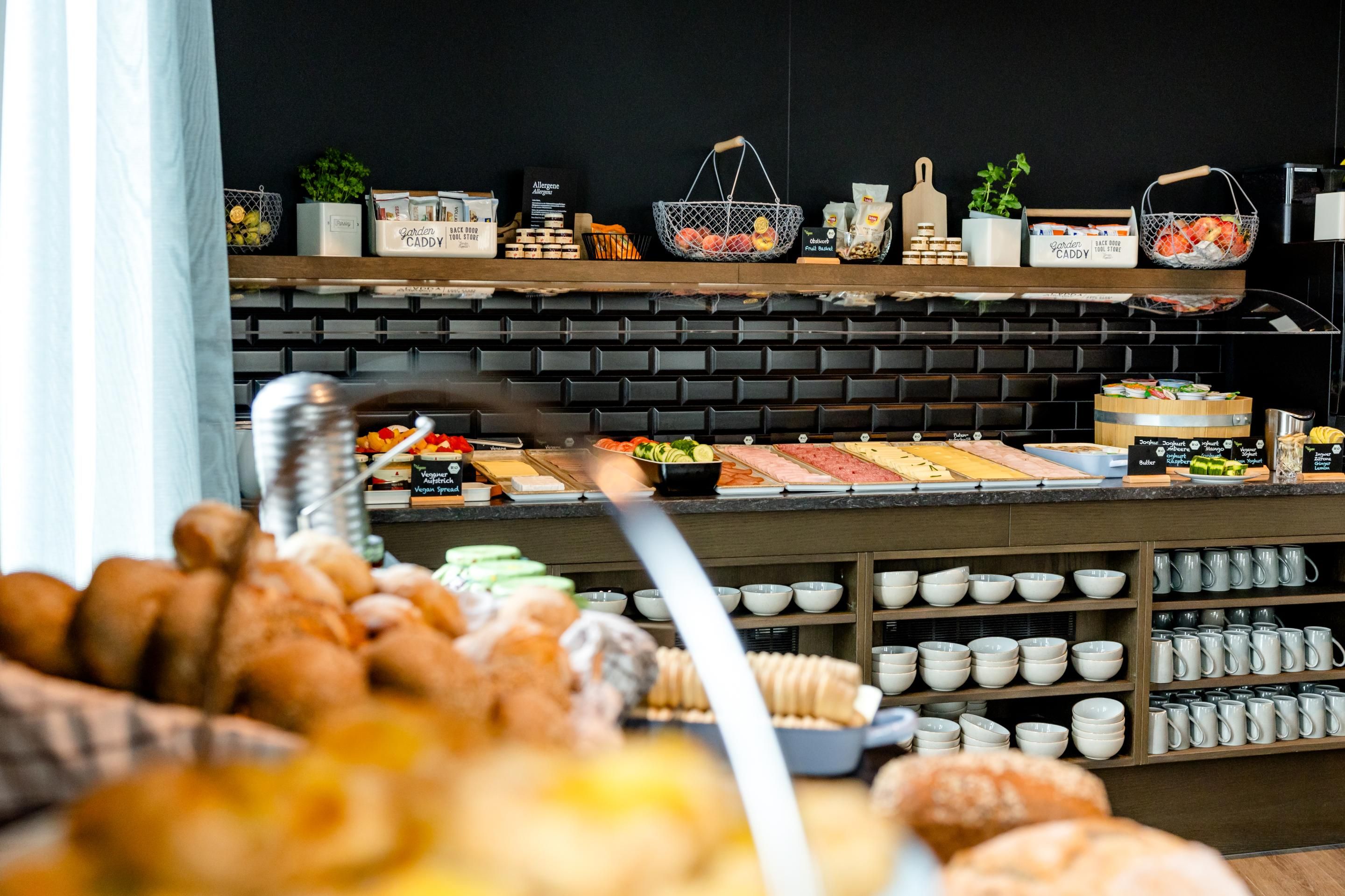 Frisches Frühstücksbuffet mit Brötchen, Croissants, Aufschnitt, Käse, Obst und Frühstückszutaten in modern gestaltetem Raum mit dunkler Wand und hellem Tageslicht.