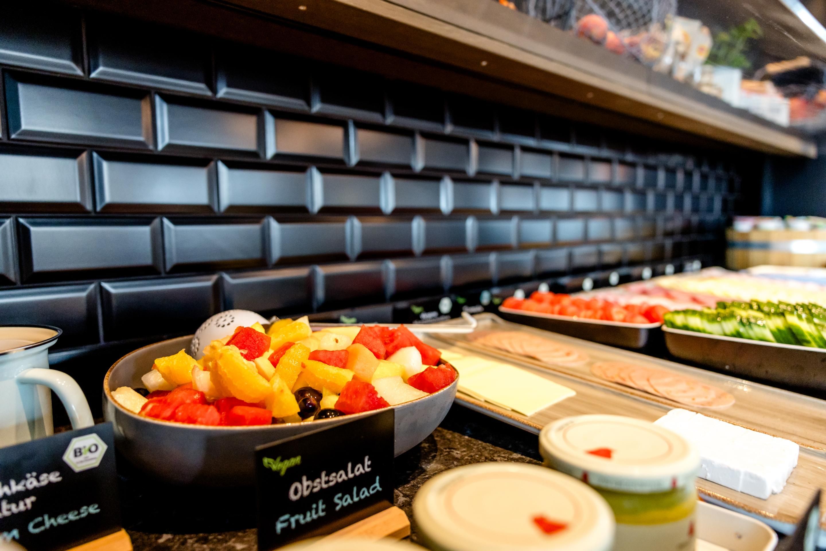 Frischer Obstsalat in einer Schale auf einem Frühstücksbuffet mit verschiedenen weiteren Speisen, präsentiert in einem modernen, einladenden Ambiente.