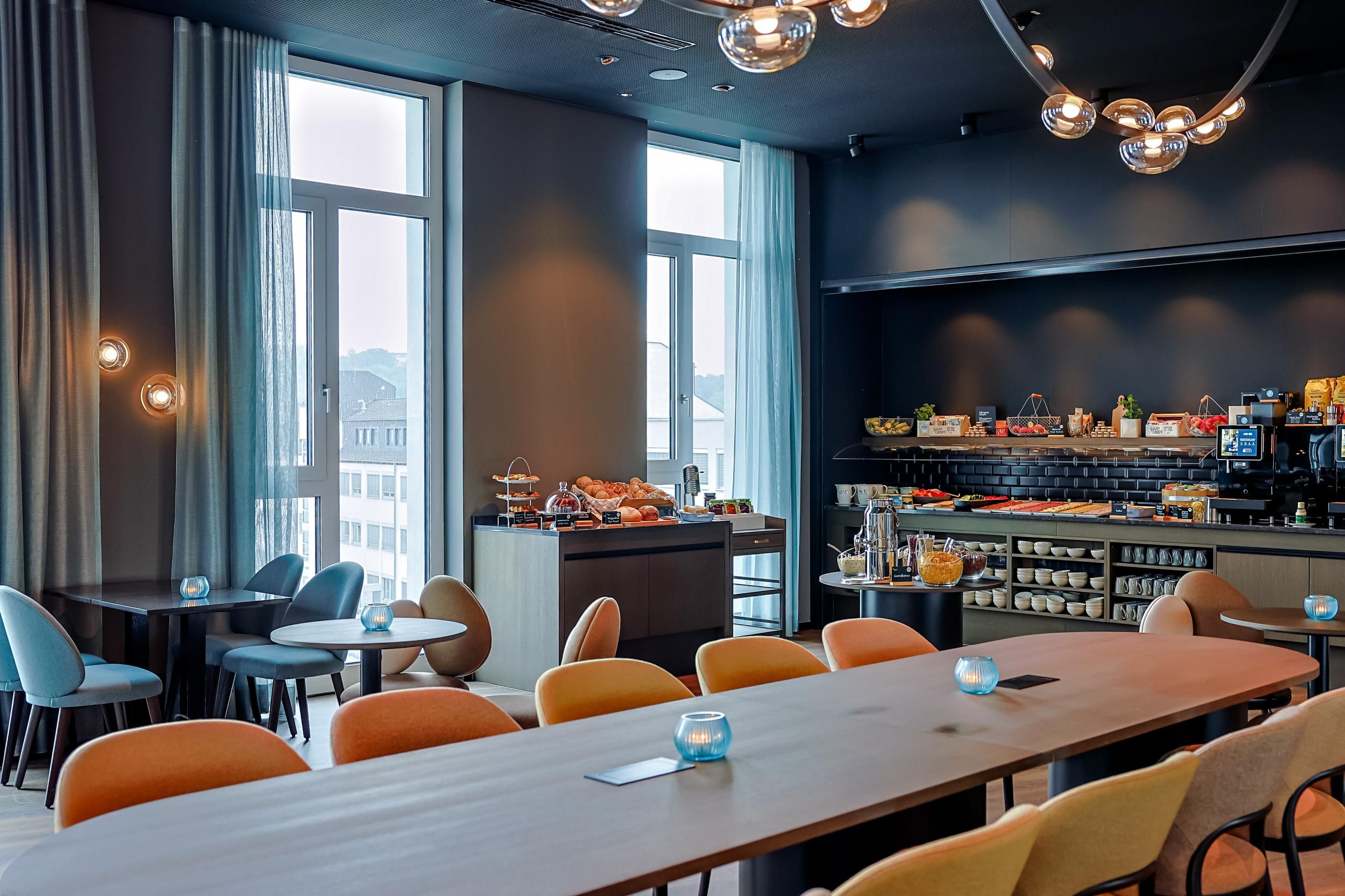 Modern eingerichteter Frühstücksraum mit großem Holztisch, bunten Stühlen und einer Buffetstation mit Getränken und Gebäck, helle Fenster für Tageslicht, warme Atmosphäre durch dezente Beleuchtung und stilvolle Dekoration.