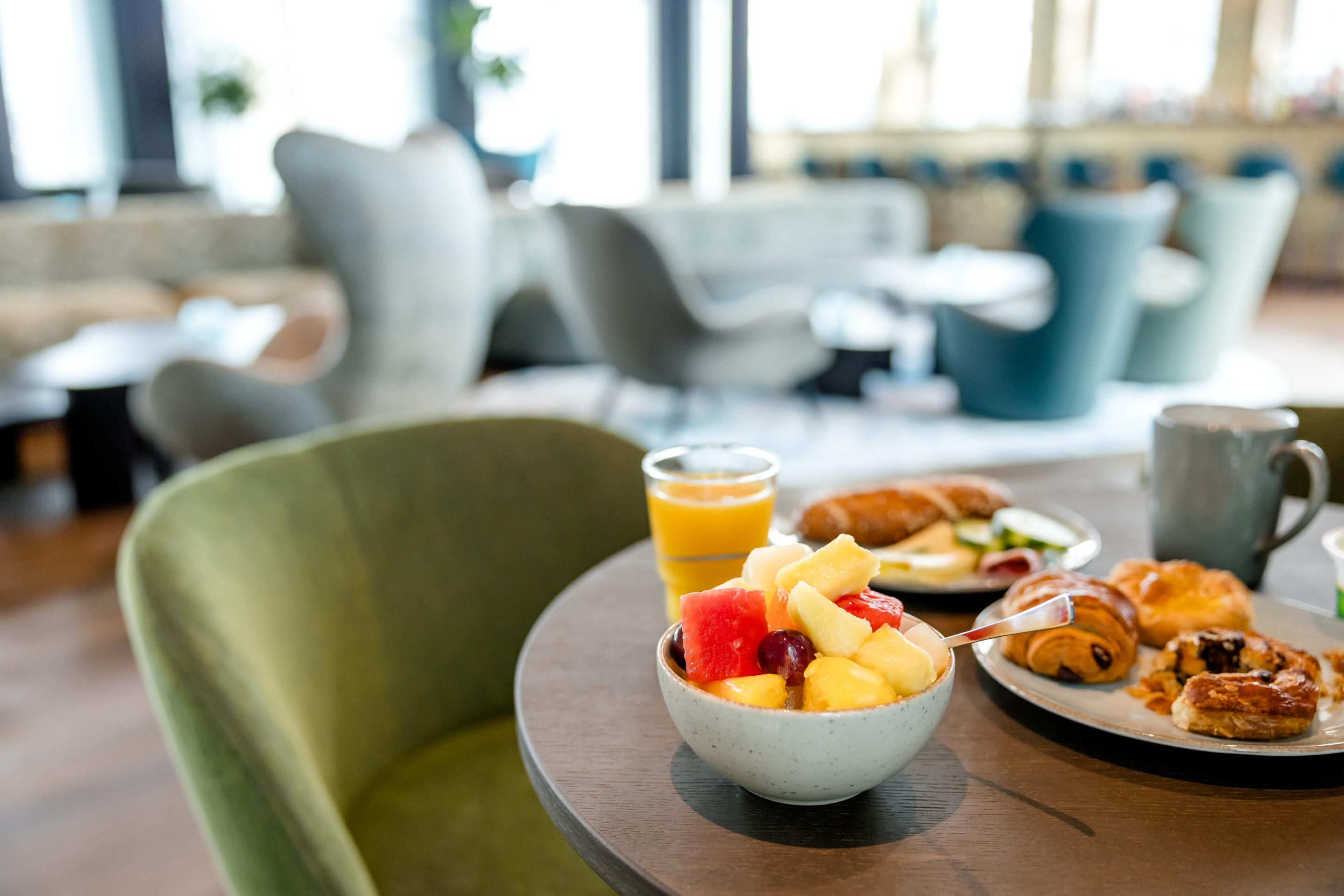 Tisch mit gesundem Frühstück: Schale frisches Obst, Glas Orangensaft, Croissants und Gebäck in einem hellen, gemütlichen Restaurant mit bequemen Sitzgelegenheiten.