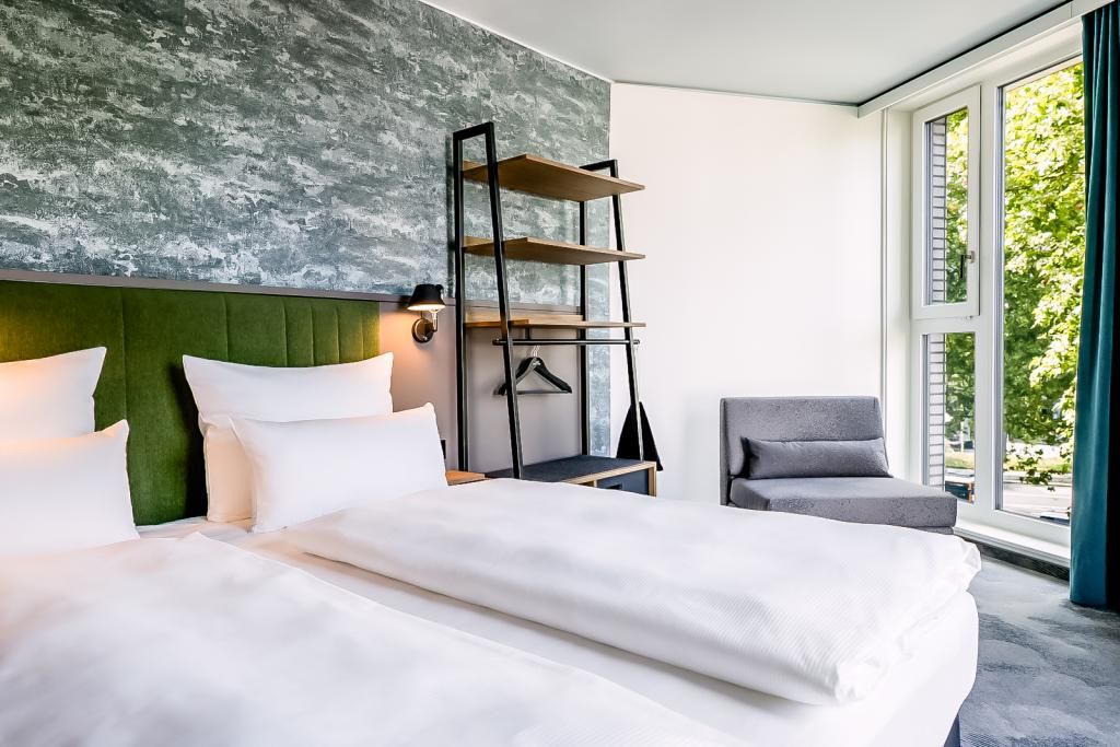 Helles Hotelzimmer mit zwei Einzelbetten, grünem Kopfteilstück und modernem Wandregal. Große Fenster bieten grünen Ausblick, daneben grauer Sessel, vermittelt ruhige, einladende Atmosphäre für entspannte Nächte.