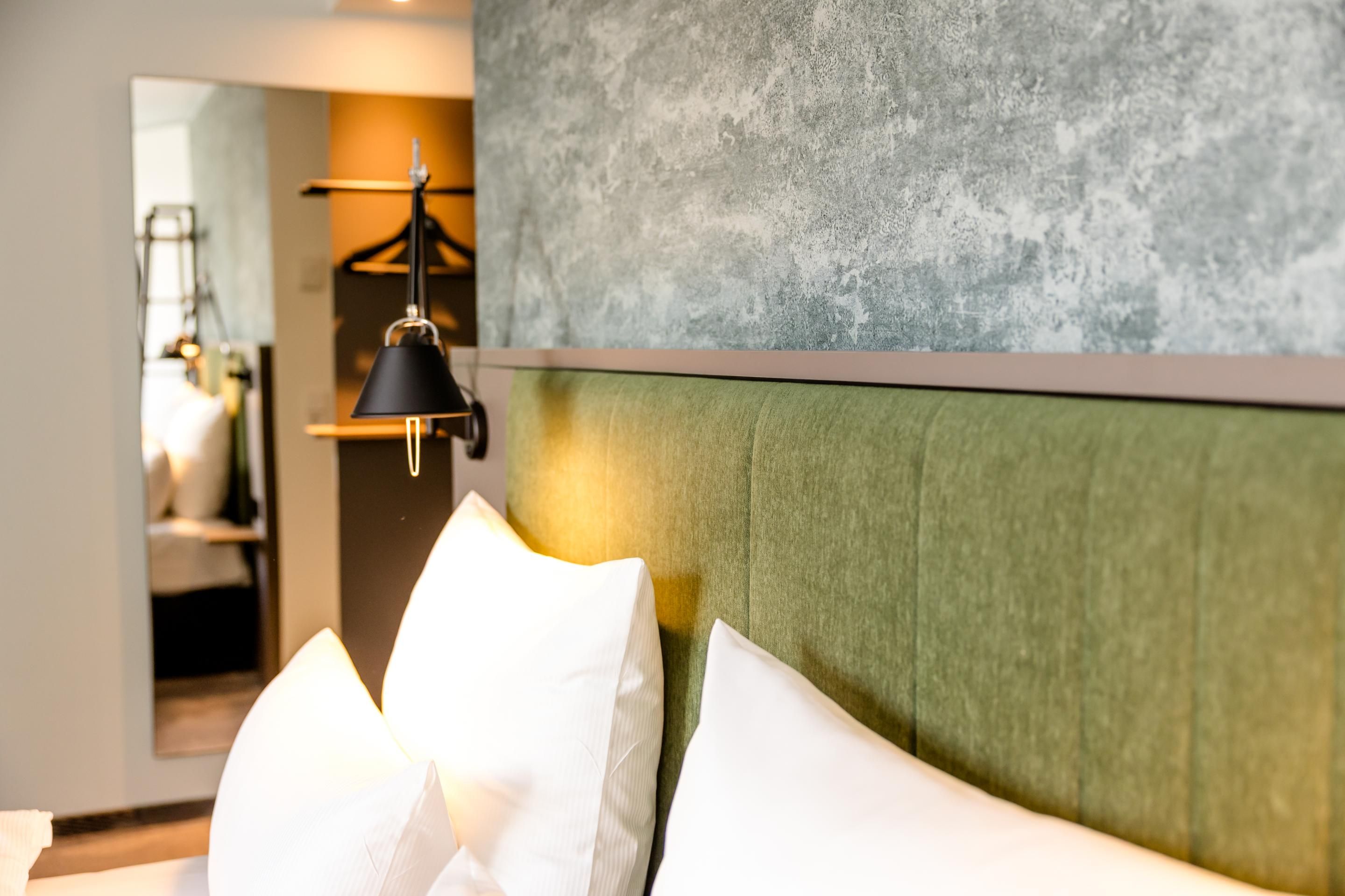 Bett mit grün gepolstertem Kopfteil, weißer Bettwäsche und Kopfkissen, moderne Wandlampe und ruhige, stilvolle Raumgestaltung für erholsamen Schlaf.